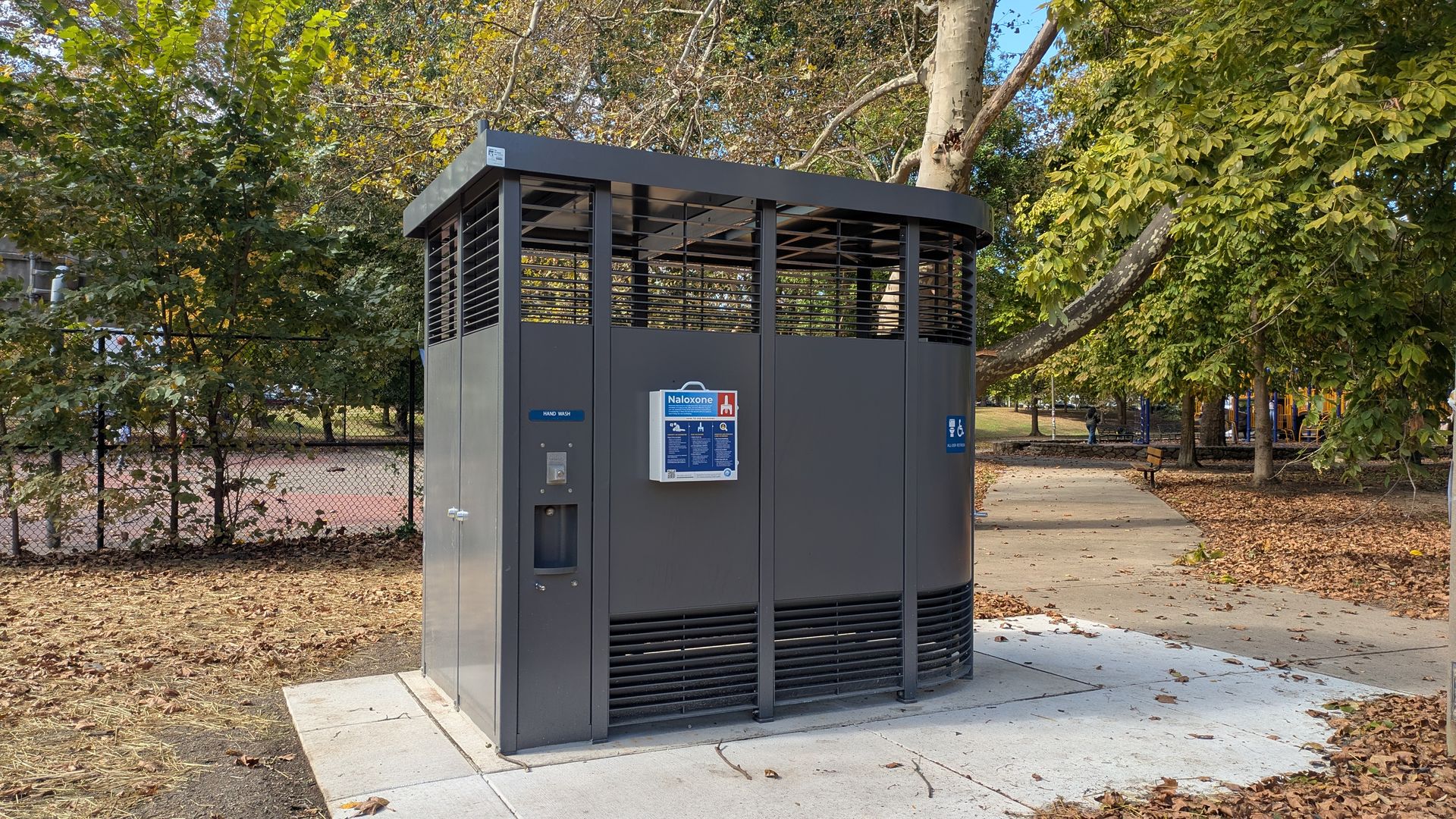 Gray public restroom with a Naloxone box and hand wash station outside, surrounded by trees with autumn leaves and a nearby playground in a park on a sunny day.