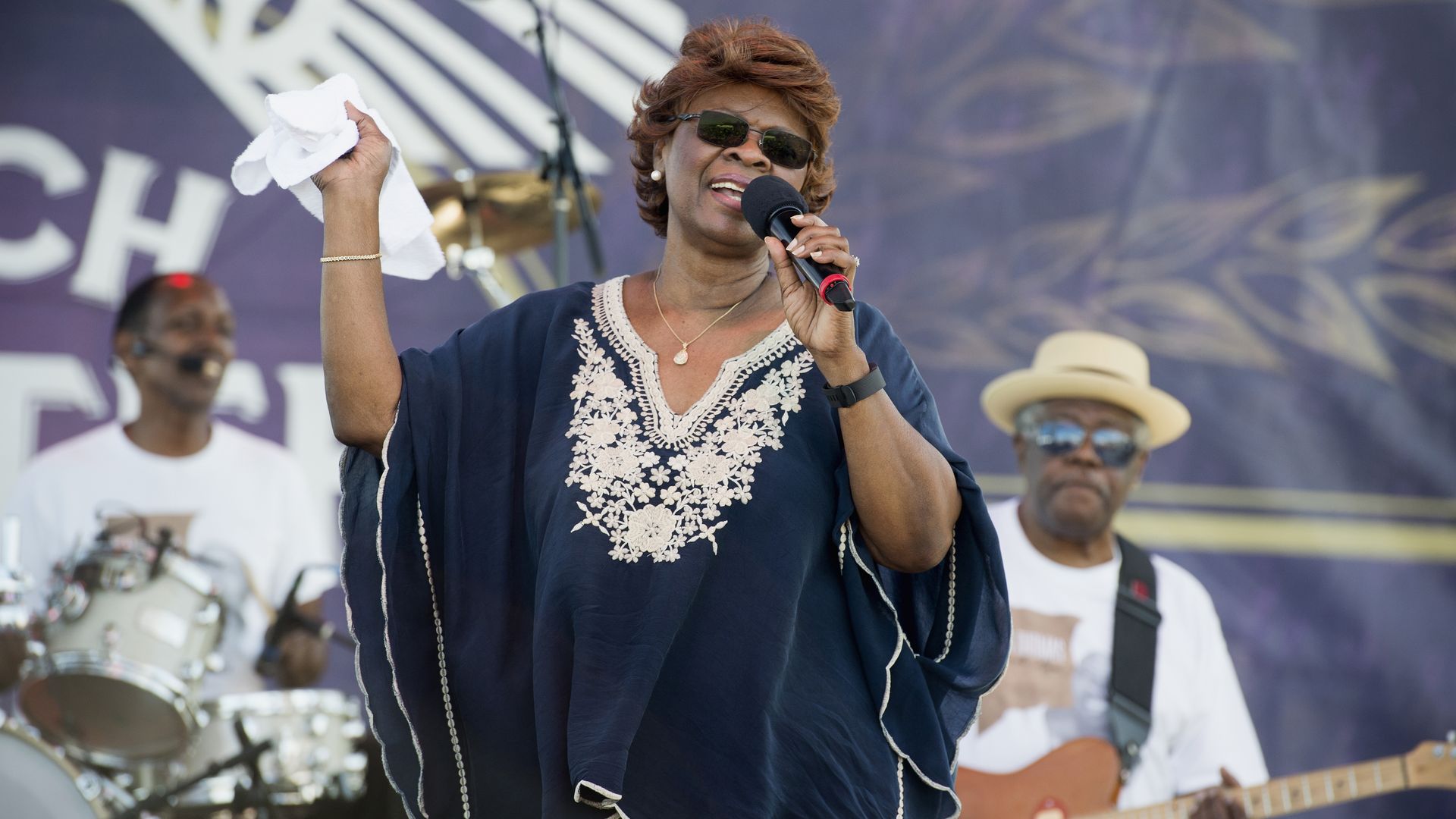 Irma Thomas performs at French Quarter Fest 2016, singing on stage with a white kerchief in one hand and wearing a dark blue caftan dress and sunglasses.