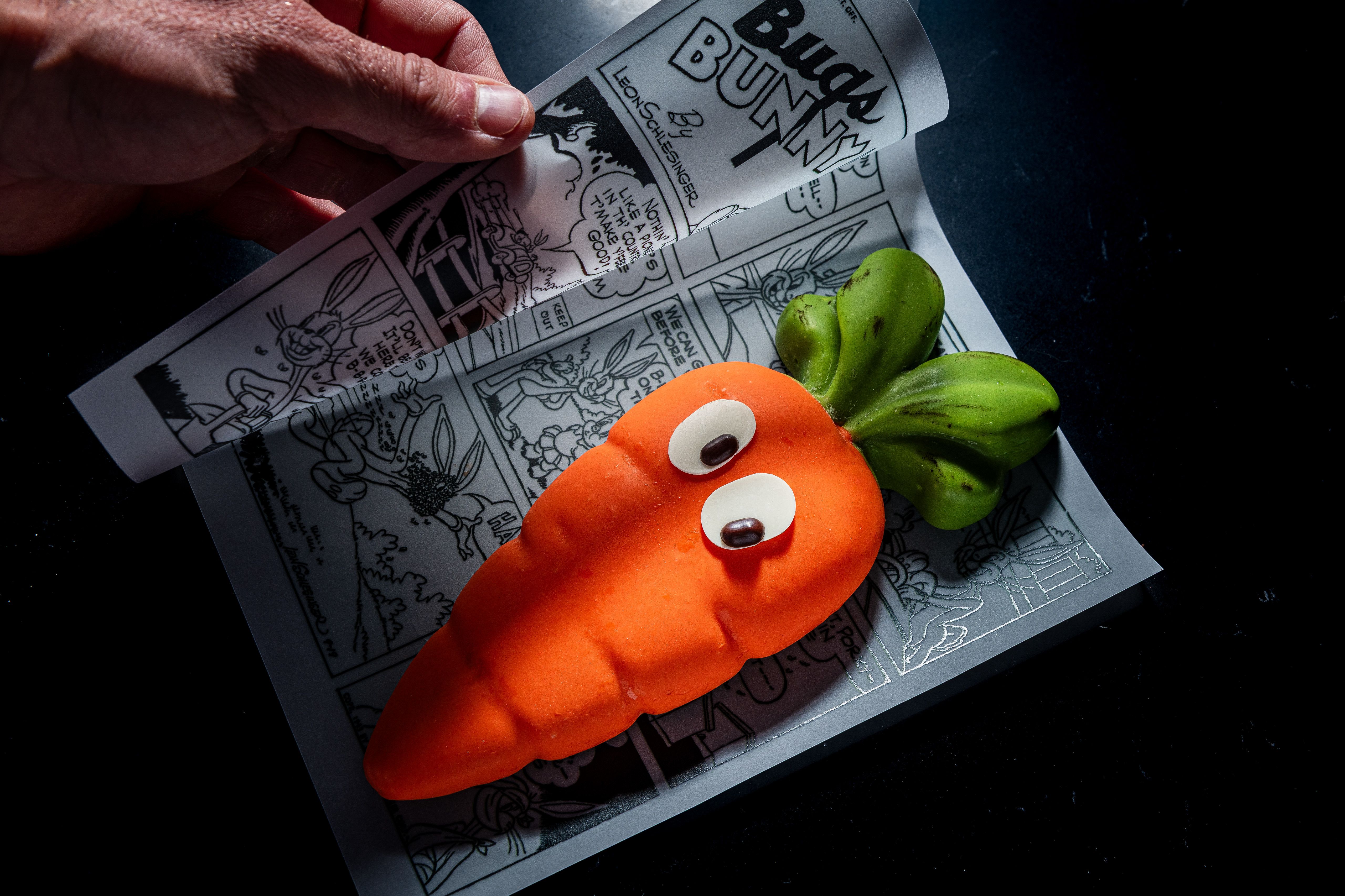 Orange carrot with a green leafy top and two white googly eyes sits on a grayscale comic-strip newspaper; a hand turns the page on a dark surface.