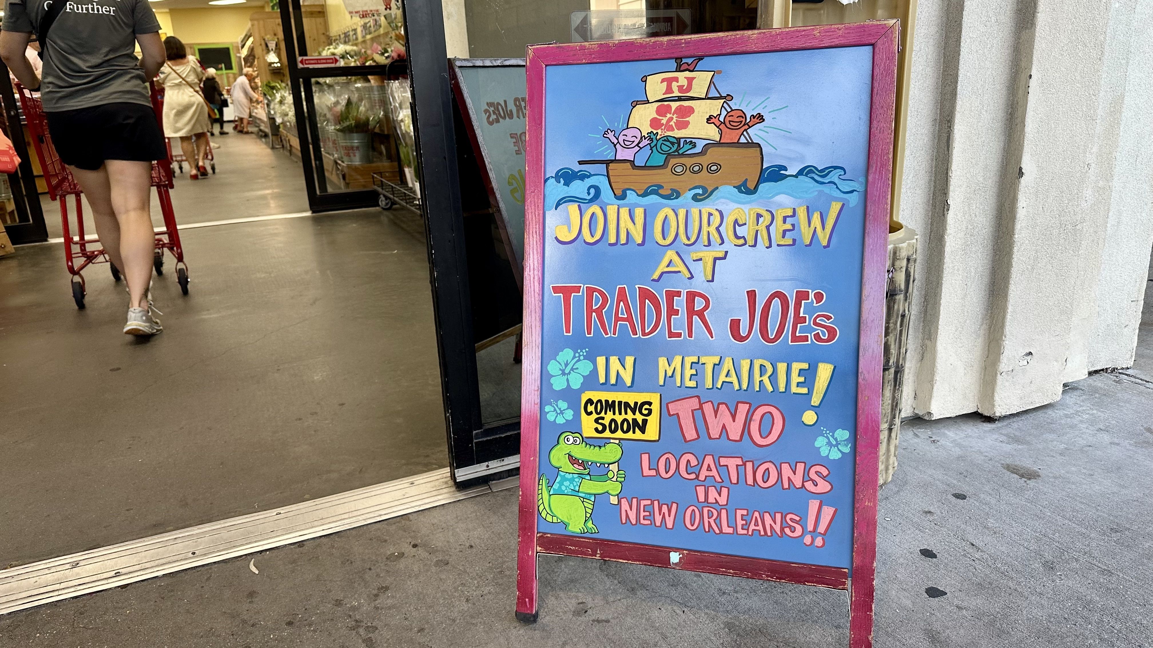 Colorful sidewalk sign outside a store reads "Join our crew at Trader Joe's in Metairie! Coming soon two locations in New Orleans!!" with cartoon boat, people, and alligator.