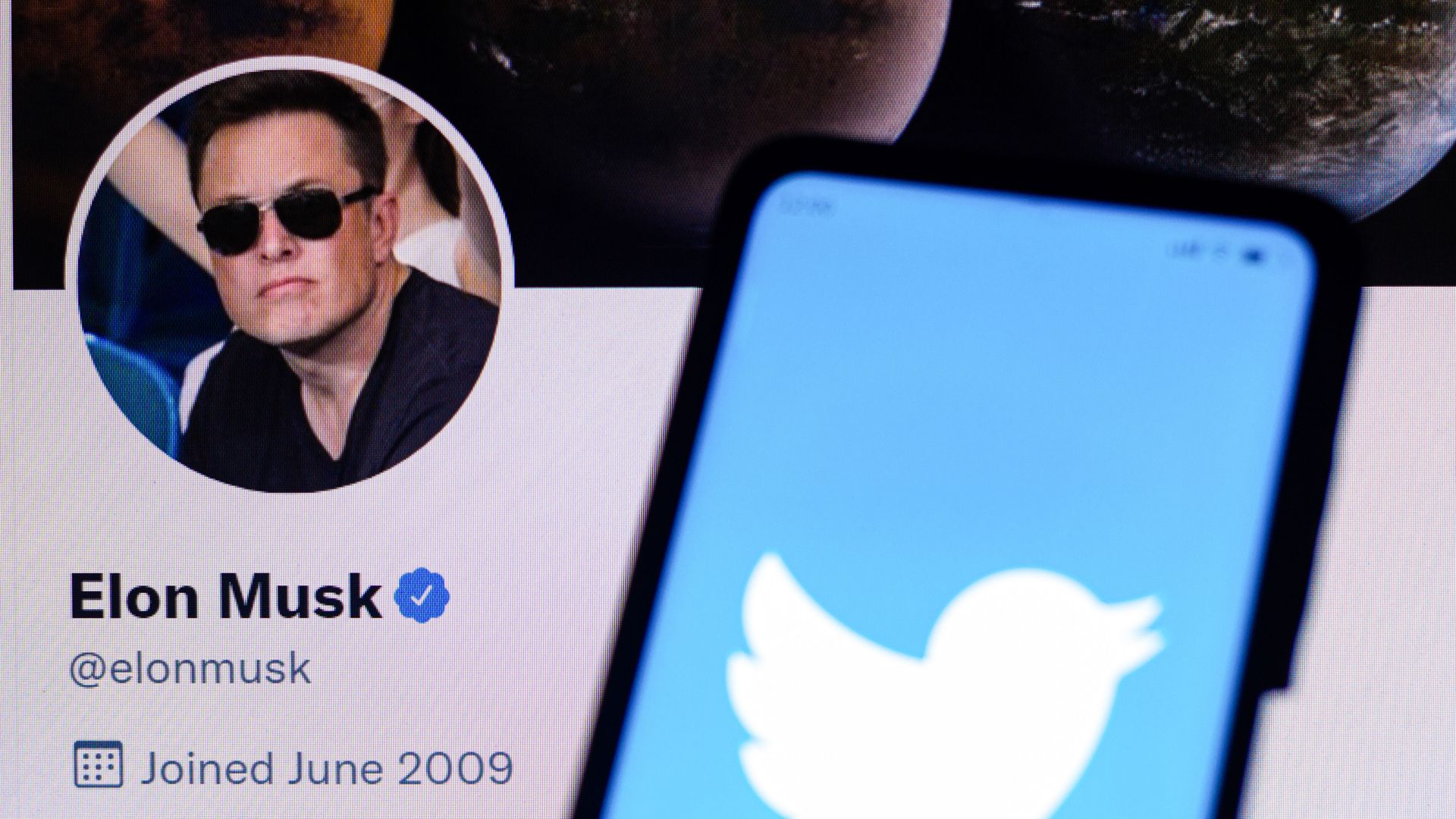 Photo of a computer showing Elon Musk's profile page and a phone showing Twitter's blue logo in front
