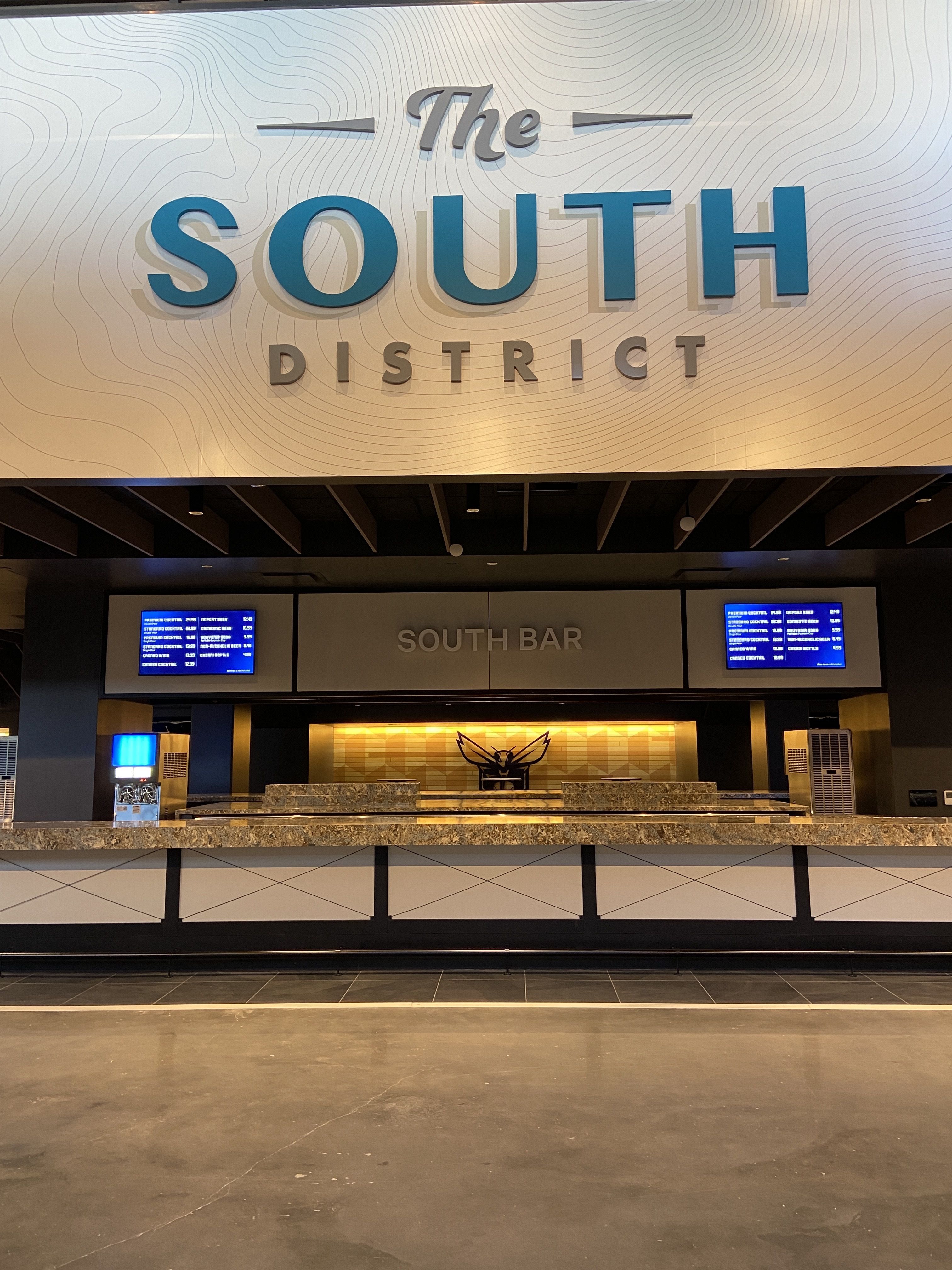 Bar area named The South District with large blue and gray sign on white patterned wall, granite countertop, two digital menu screens, and a backlit yellow wall with a black bee logo.