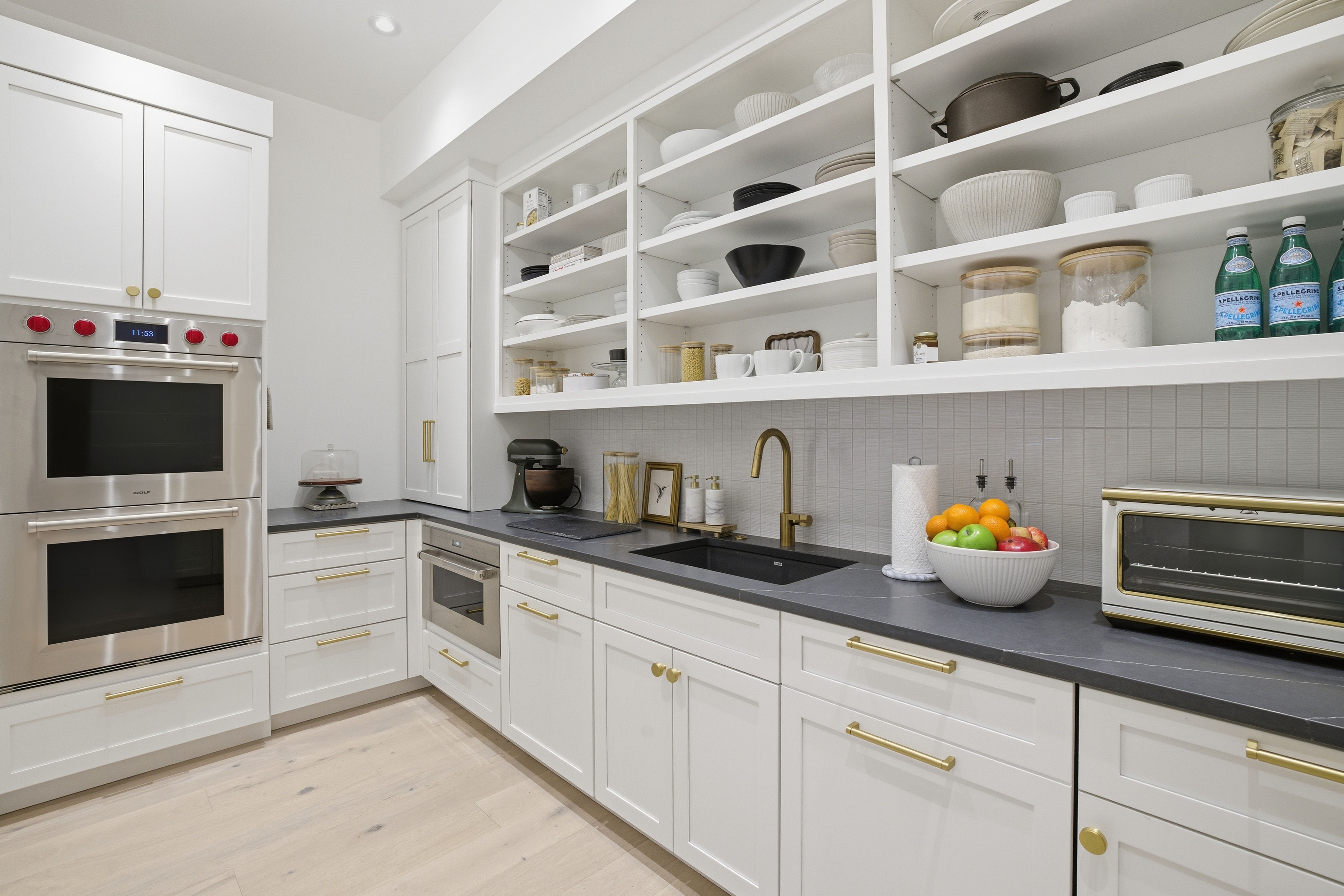 Modern white kitchen with black countertop, brass handles, double stainless steel oven with red knobs, open shelves with dishes, fruit bowl, and gold faucet over black sink.