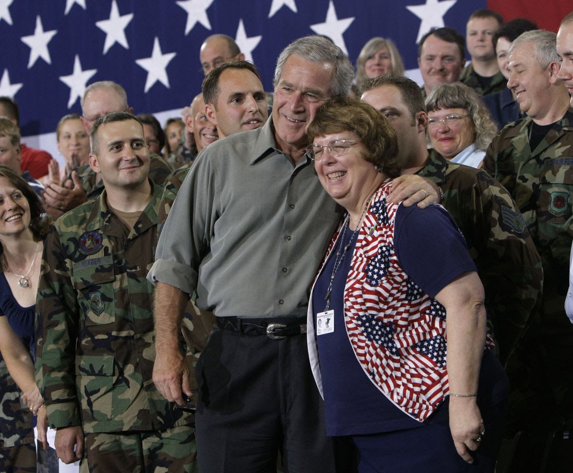President George W. Bush surrounded by troops and a woman and an America flag vest.