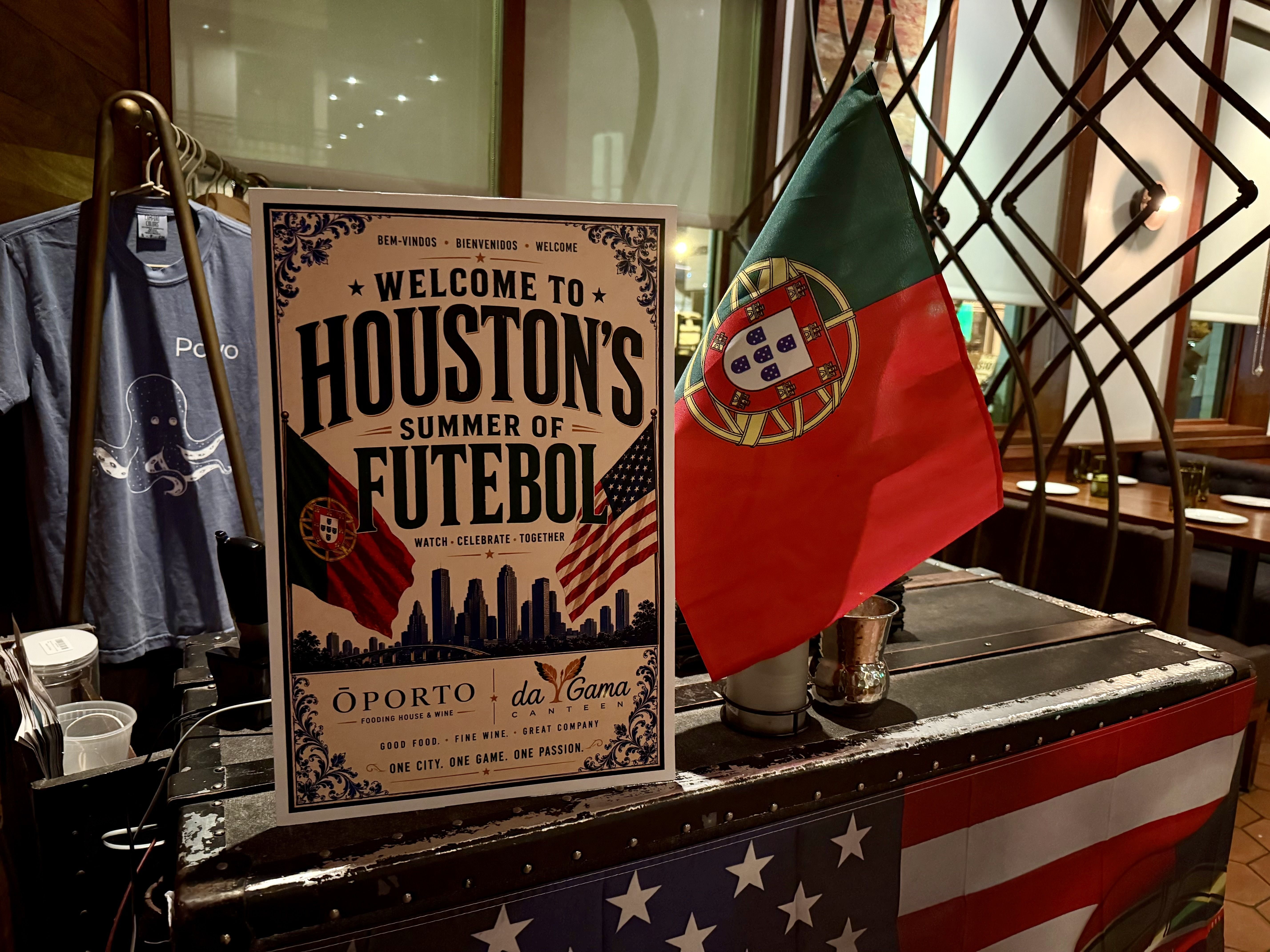 Indoor display featuring a vintage poster reading "Welcome to Houston's Futebol" beside a green-red Portuguese flag; a blue T-shirt on a hanger to the left and a lattice divider in the background.