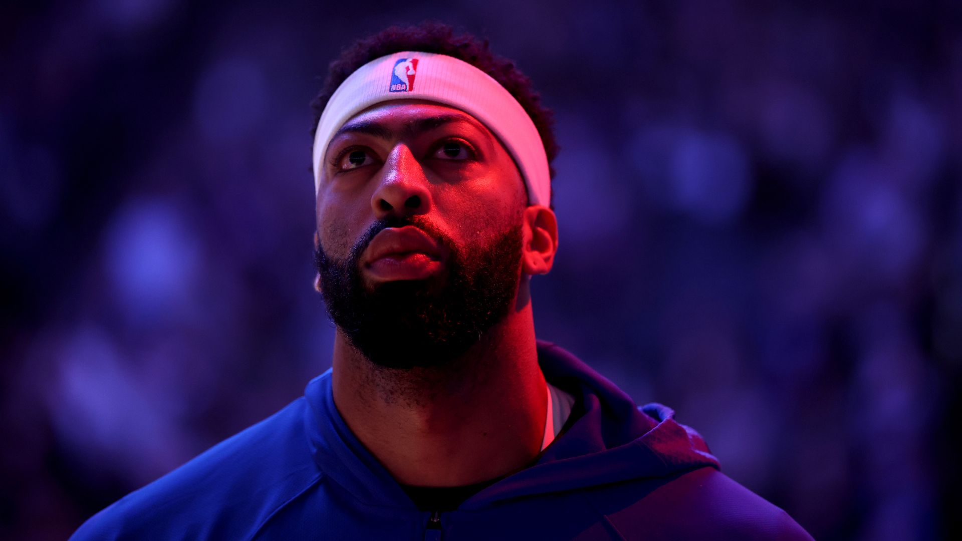 A close-up photo of NBA player Anthony Davis wearing a white NBA headband and a blue hoodie against a darkened arena background