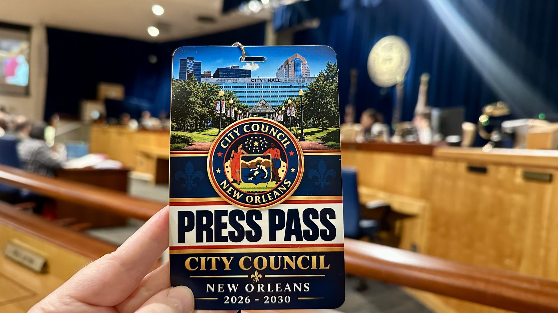 Close-up of a New Orleans City Council press pass featuring the City Hall image, a colorful seal, and bold PRESS PASS text, held in a blurred council chamber.
