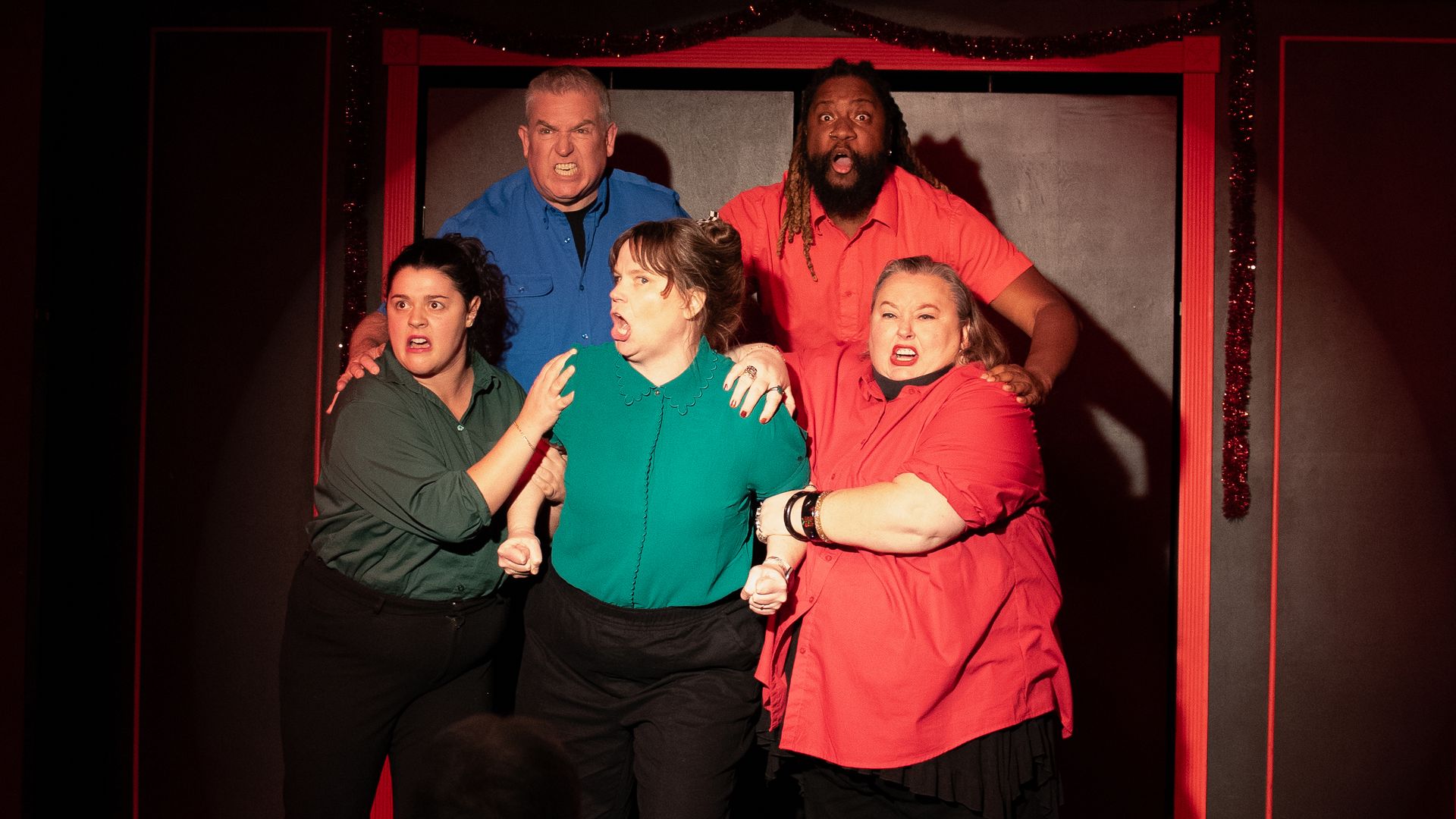 Five actors on stage dressed in colorful shirts with tense, surprised expressions under a disco ball and string lights, framed by red and black stage set.