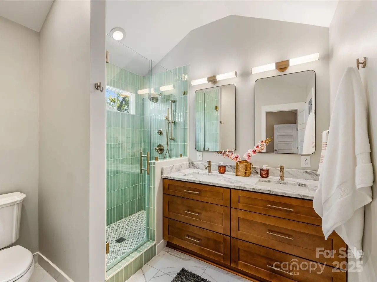 Modern bathroom with a glass shower featuring green vertical tiles, dual sinks with marble countertop, wooden vanity, two rectangular mirrors, bright white walls, and a white towel hanging.