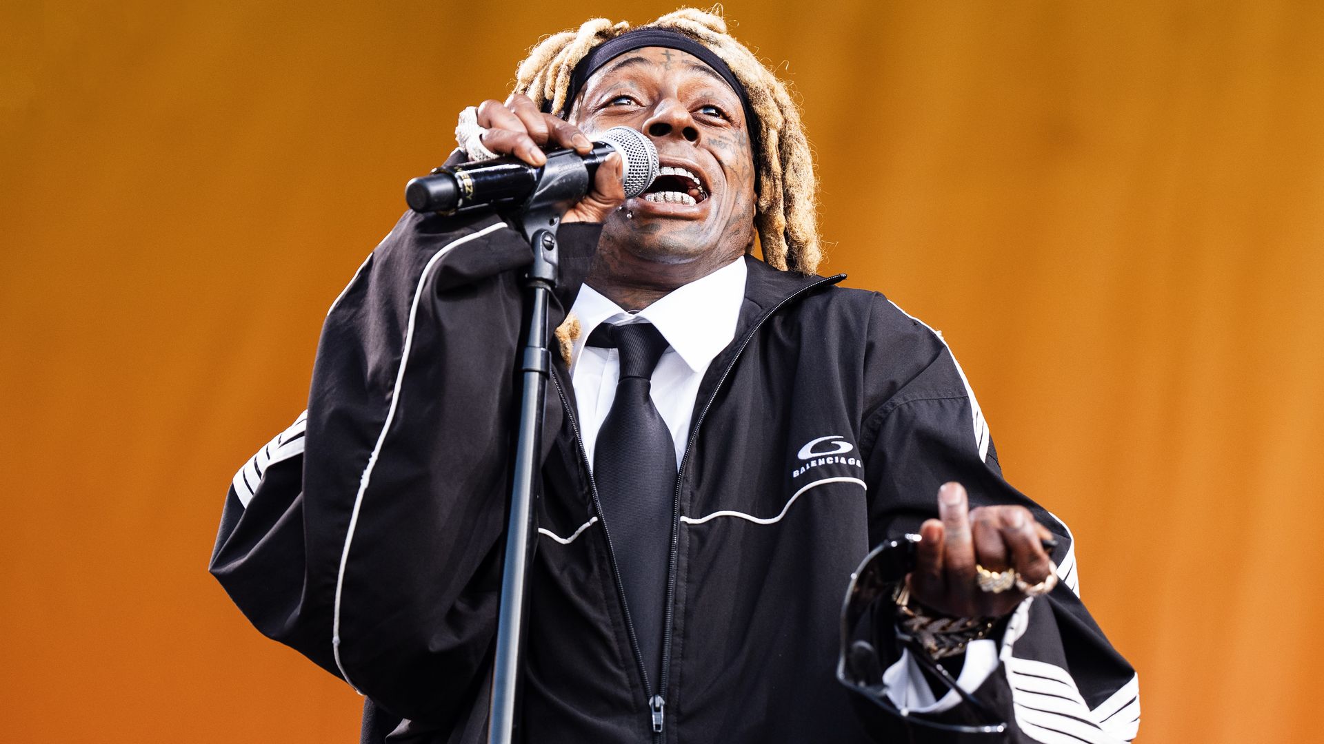 Lil Wayne is photographed wearing a black and white outfit and performing on a Jazz Fest stage. He's mid-verse in the photo as he pulls a mic toward his mouth.
