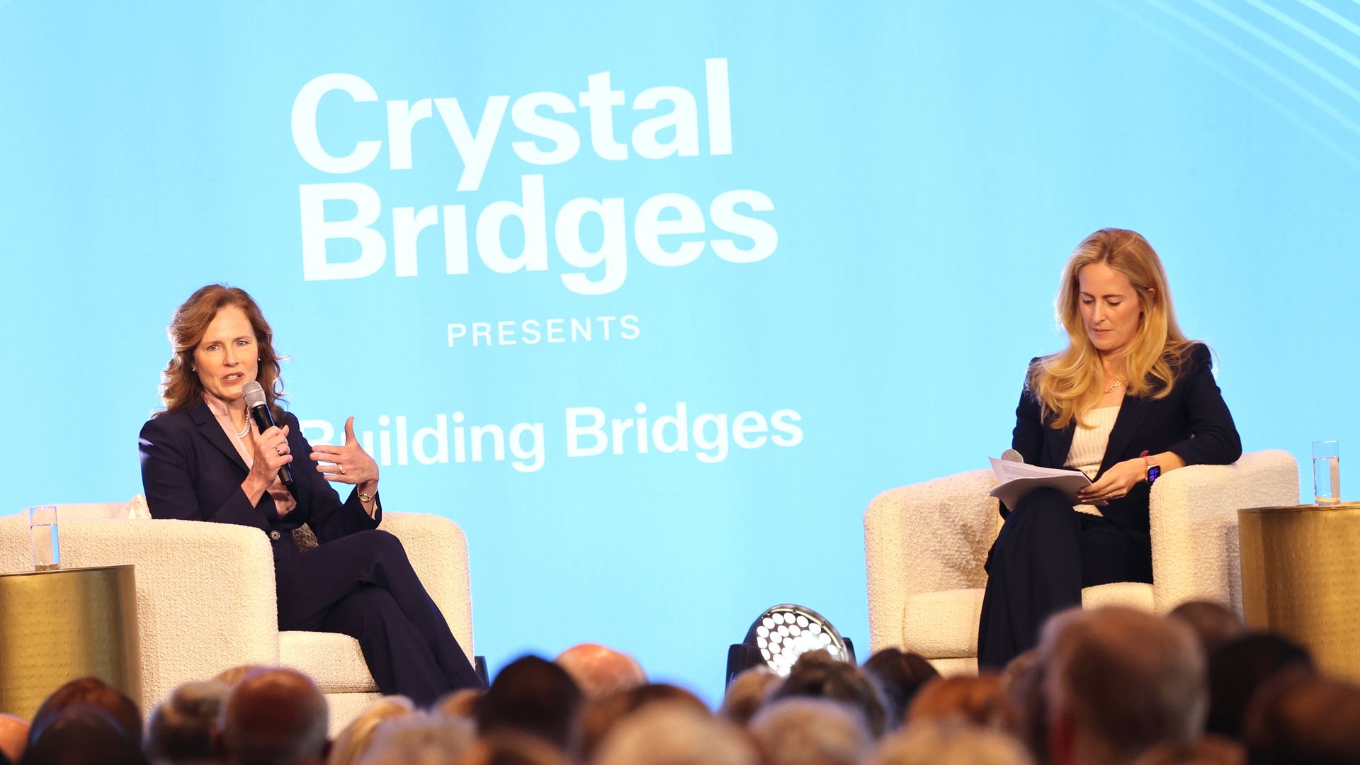 Two women sit on a stage in cream armchairs against a blue backdrop reading Crystal Bridges Presents Building Bridges; one speaks into a microphone while the other reviews papers as the audience watches.