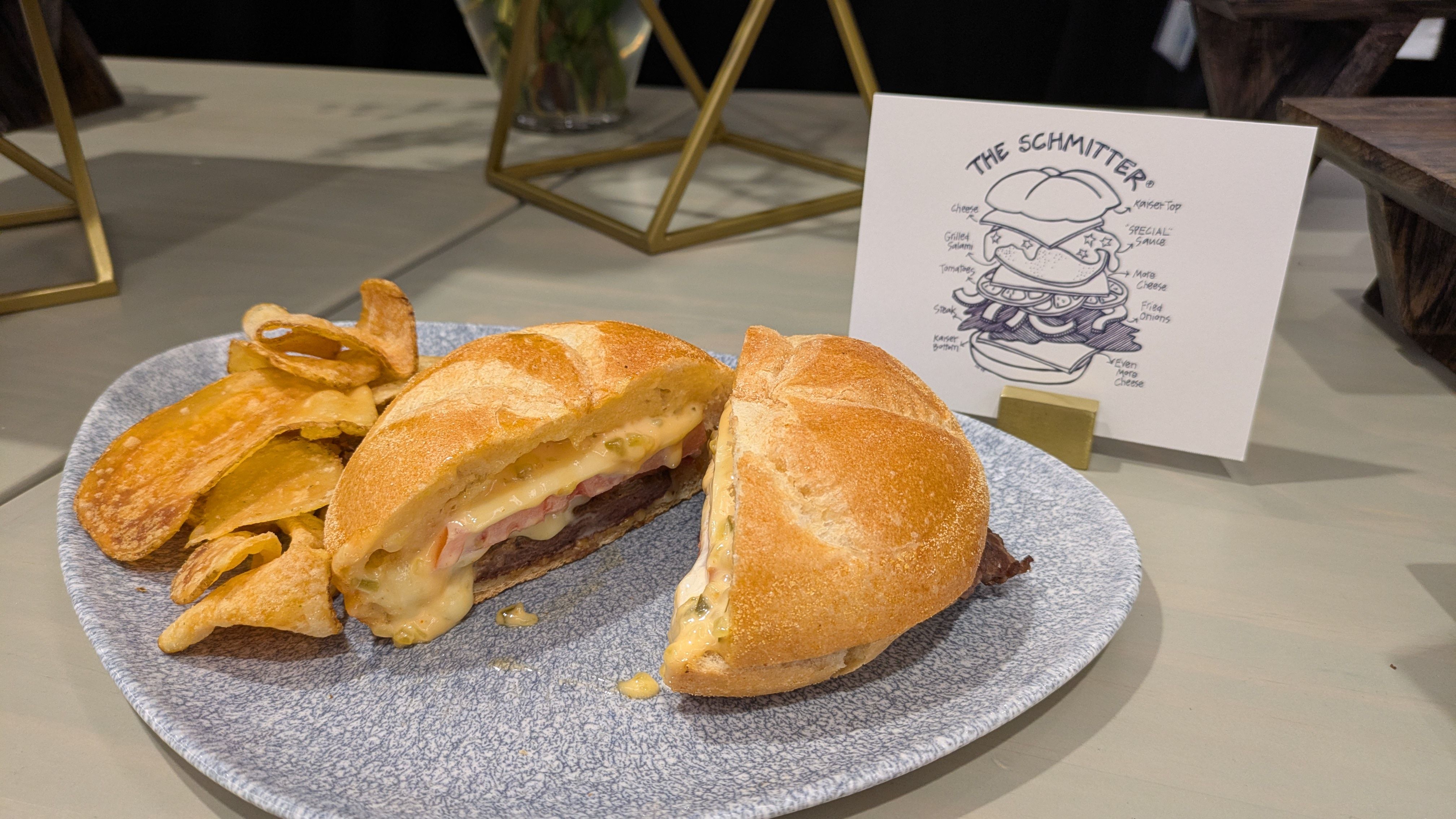 Blue speckled plate holds a sliced burger with melted cheese, onions, and tomato; chips lie nearby. A white card with a drawing of "The Schmitter" rests on the table, with gold furniture in the background.