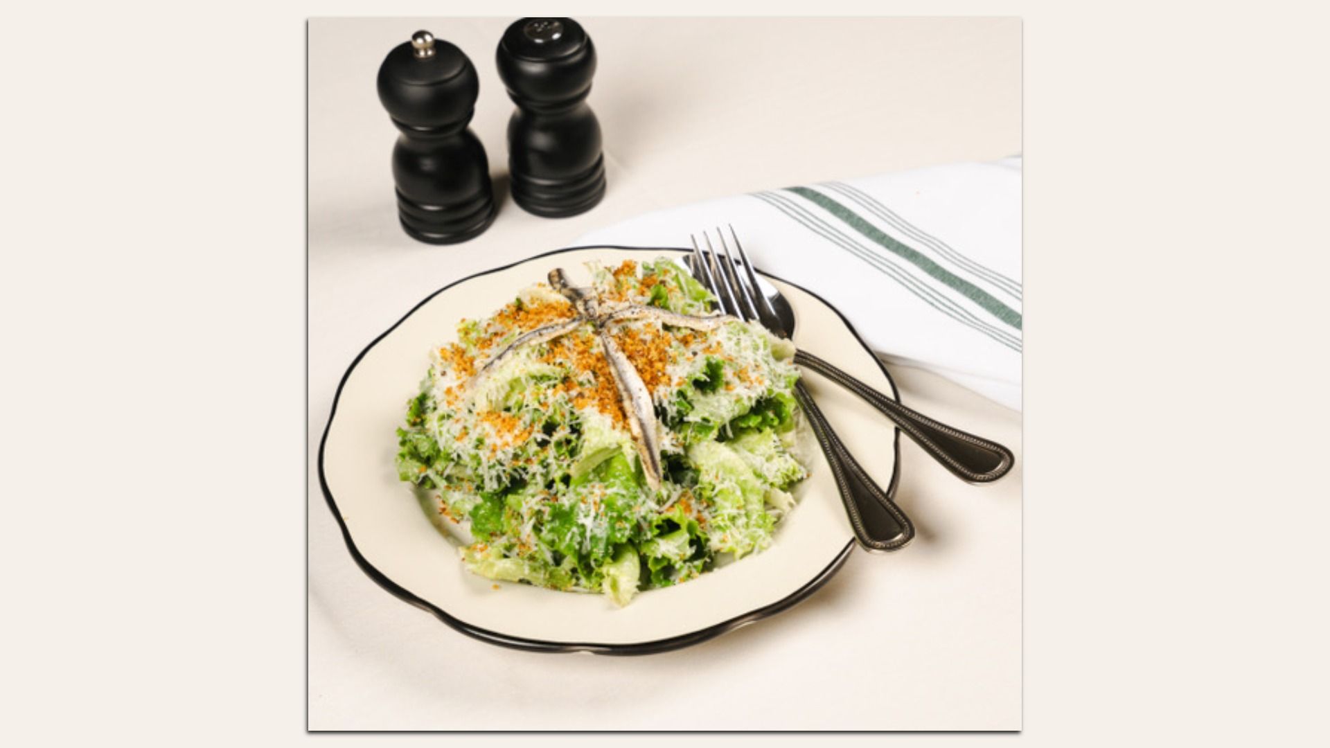 Plate of mixed greens salad with grated cheese and orange crumbs, two forks resting on the rim, and black pepper grinders in the background.