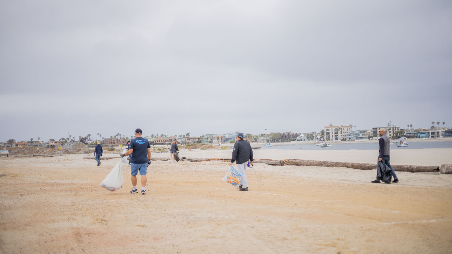 Overcast beach where several people pick up litter, carrying large bags. A log lies near the water; distant houses line the shore and a few boats float in the bay.