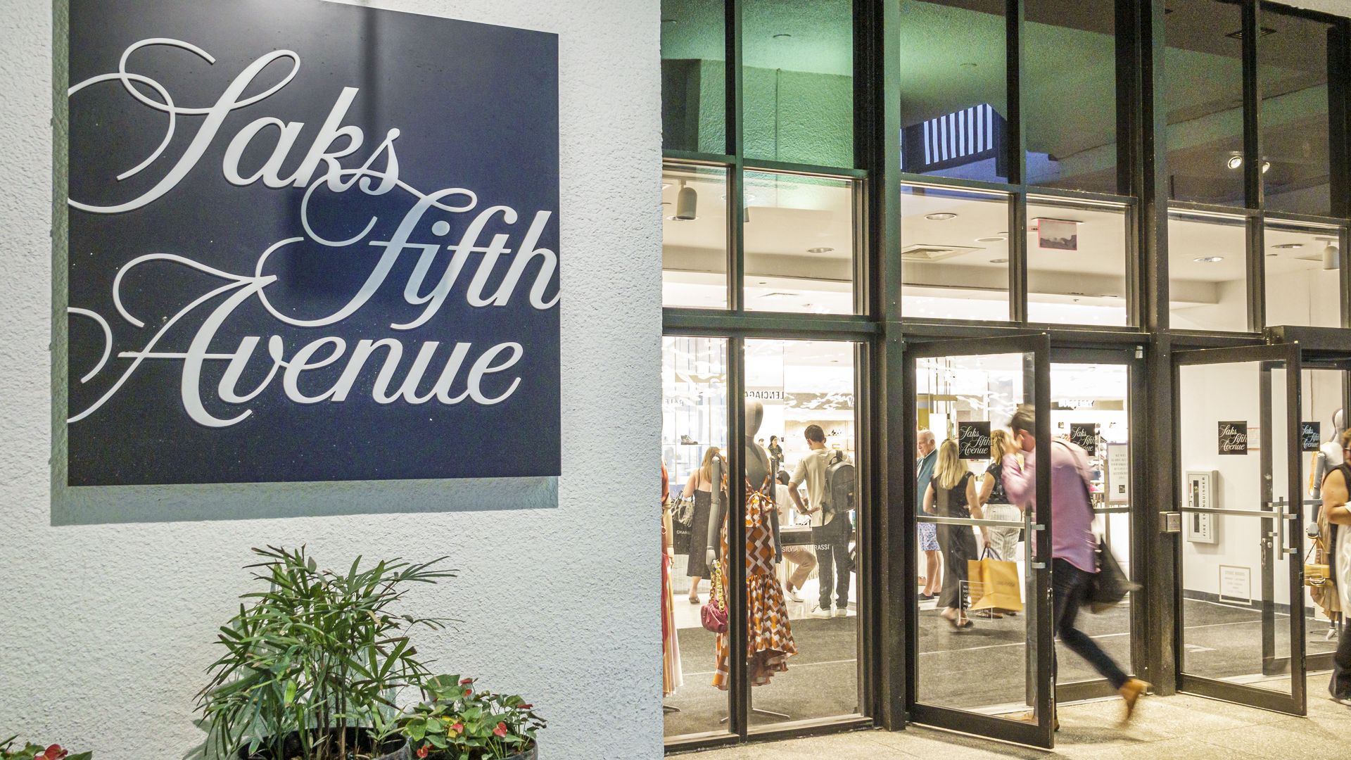 Entrance of Saks Fifth Avenue store with glass doors, people walking in and out, and black sign with white cursive letters. Potted plants near the sign on a textured white wall.