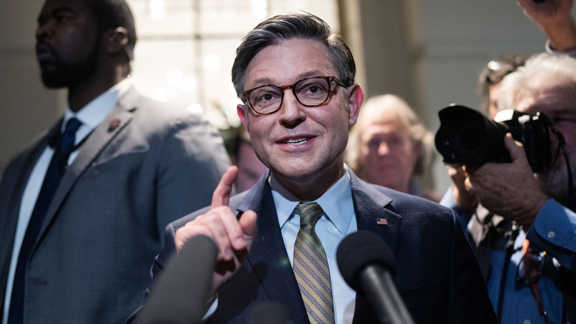 House Speaker Mike Johnson, wearing a blue suit and speaking into microphones while flanked by press and staff.