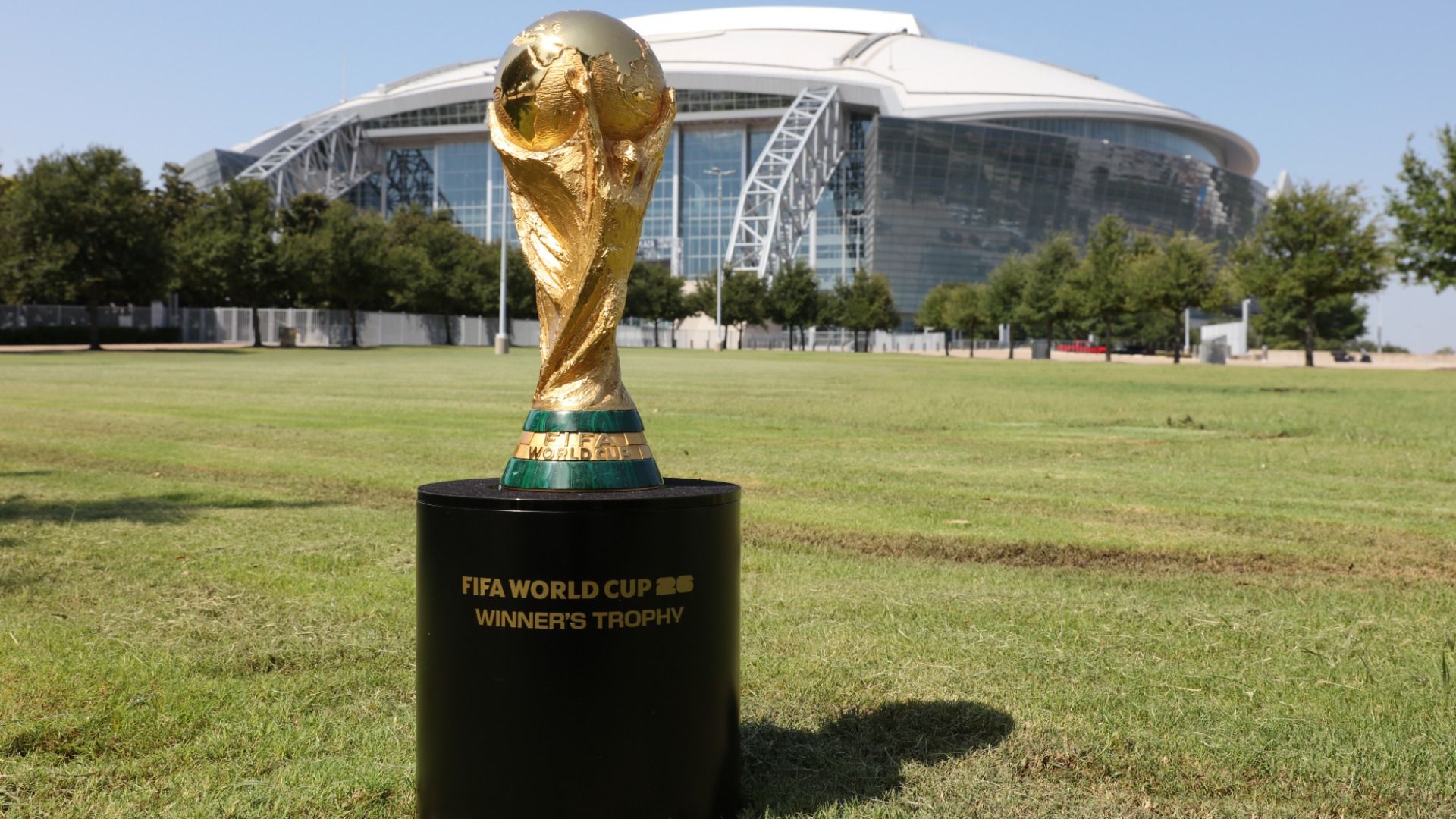 Golden FIFA World Cup winner's trophy placed on a black pedestal on green grass with a large modern stadium and trees in the background under a clear blue sky.