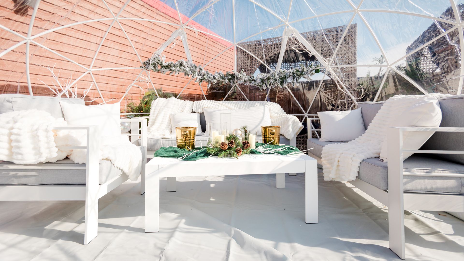 Cozy outdoor seating area inside a transparent dome with white-framed gray sofas, white blankets, pillows, and a white table decorated with candles, pinecones, and greenery under sunshine.
