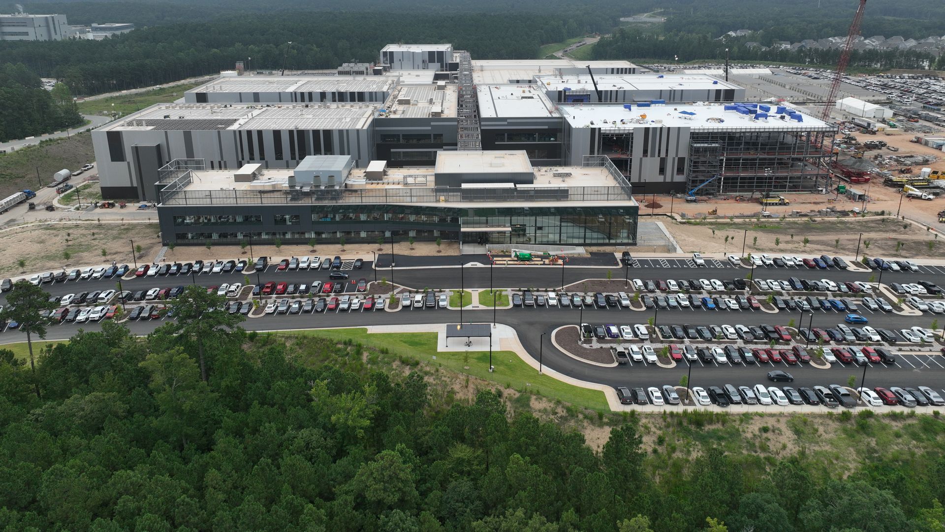 A large industrial complex with gray buildings under overcast sky, surrounded by dense green forest, parking lots full of cars, and a crane on the right side near construction.