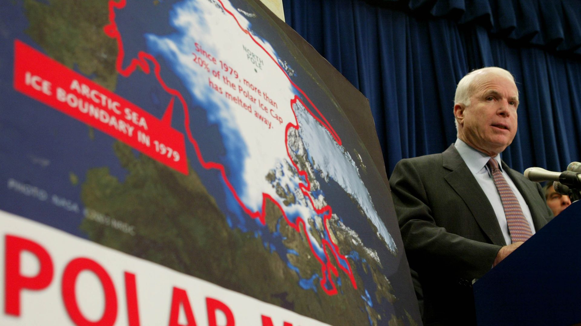 John McCain speaking next to a climate change poster