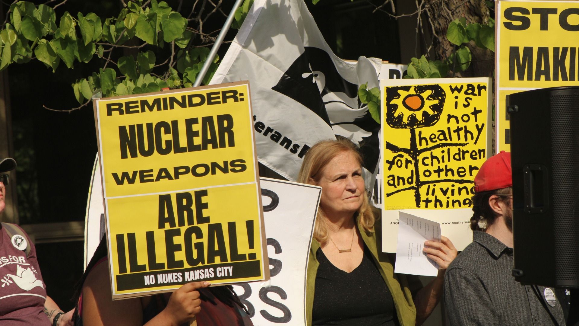 Protesters hold bright yellow signs reading "REMINDER: NUCLEAR WEAPONS ARE ILLEGAL!" and a poster about war's harm to children; a blonde woman stands among banners and green leaves.