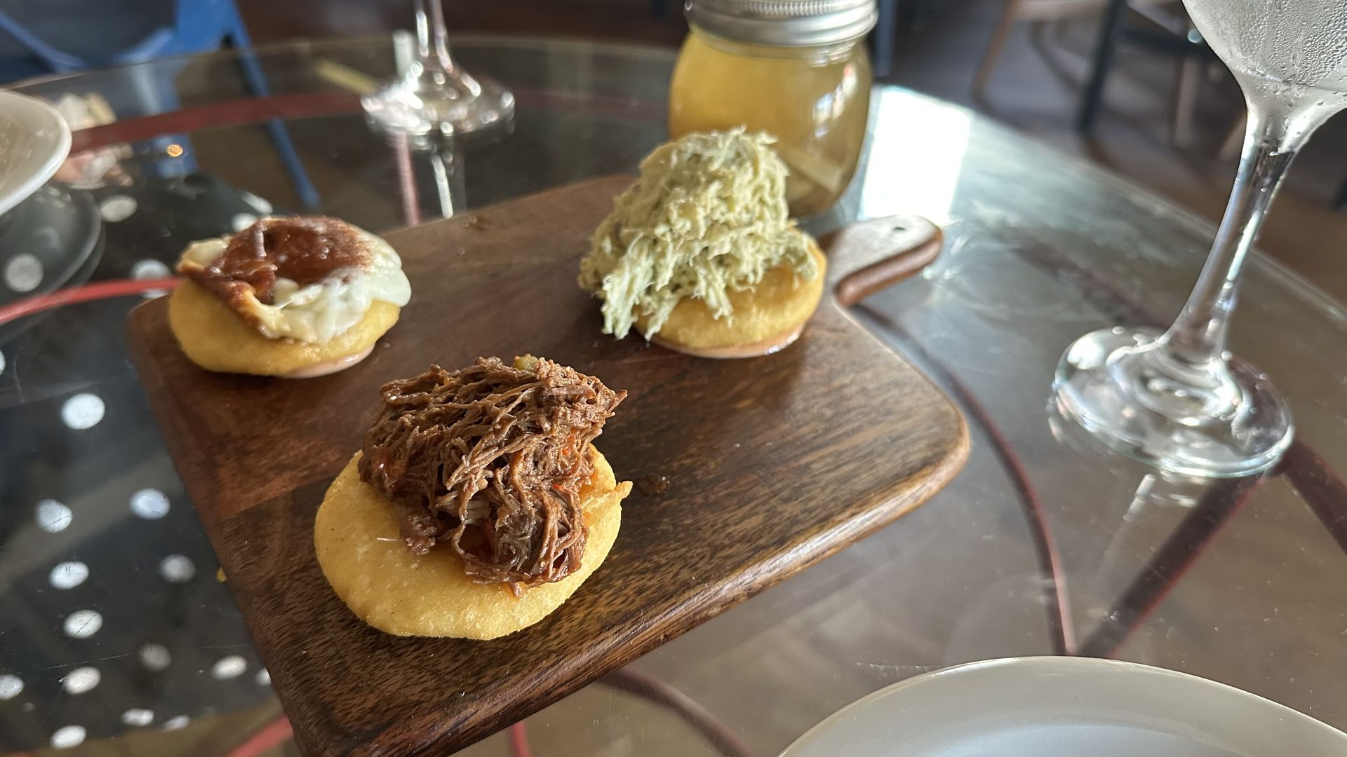 A photo of three arepas served on a wooden cutting board. A jar of yellowish hot sauce sits behind it.