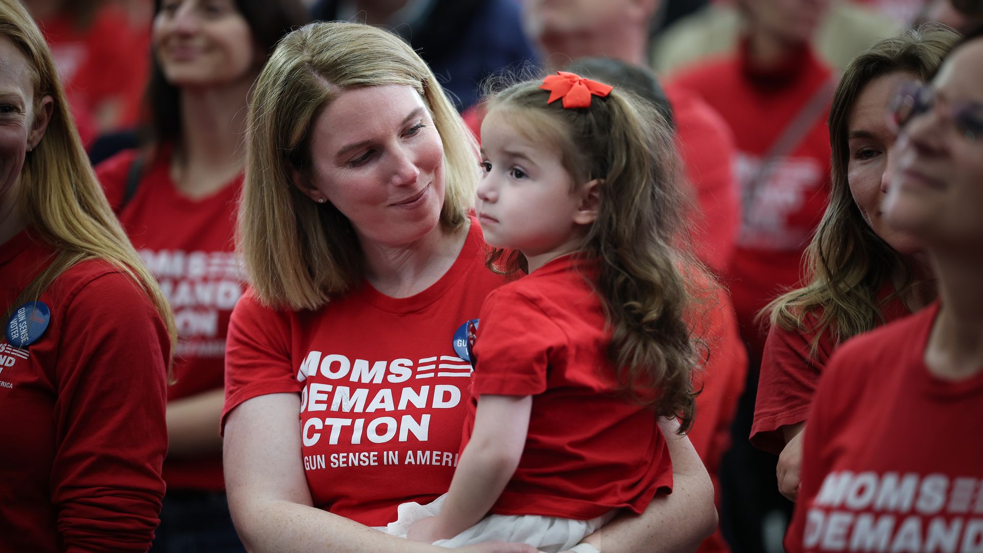 A blonde woman holds a young girl in matching red shirts at a crowded rally. The girl has an orange bow in her hair; both look at each other. Their shirts read "MOMS DEMAND ACTION".