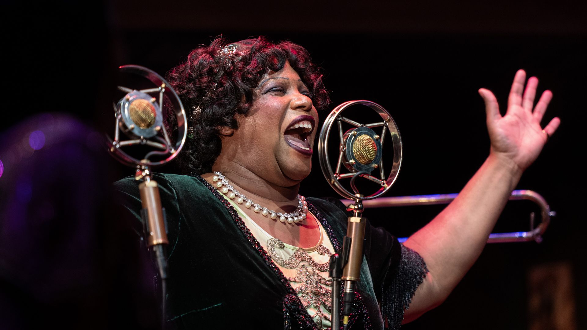 A smiling Black female singer with curly hair wears a pearl necklace and dark attire, singing into two vintage microphones on a dark stage, her right hand raised in the air.