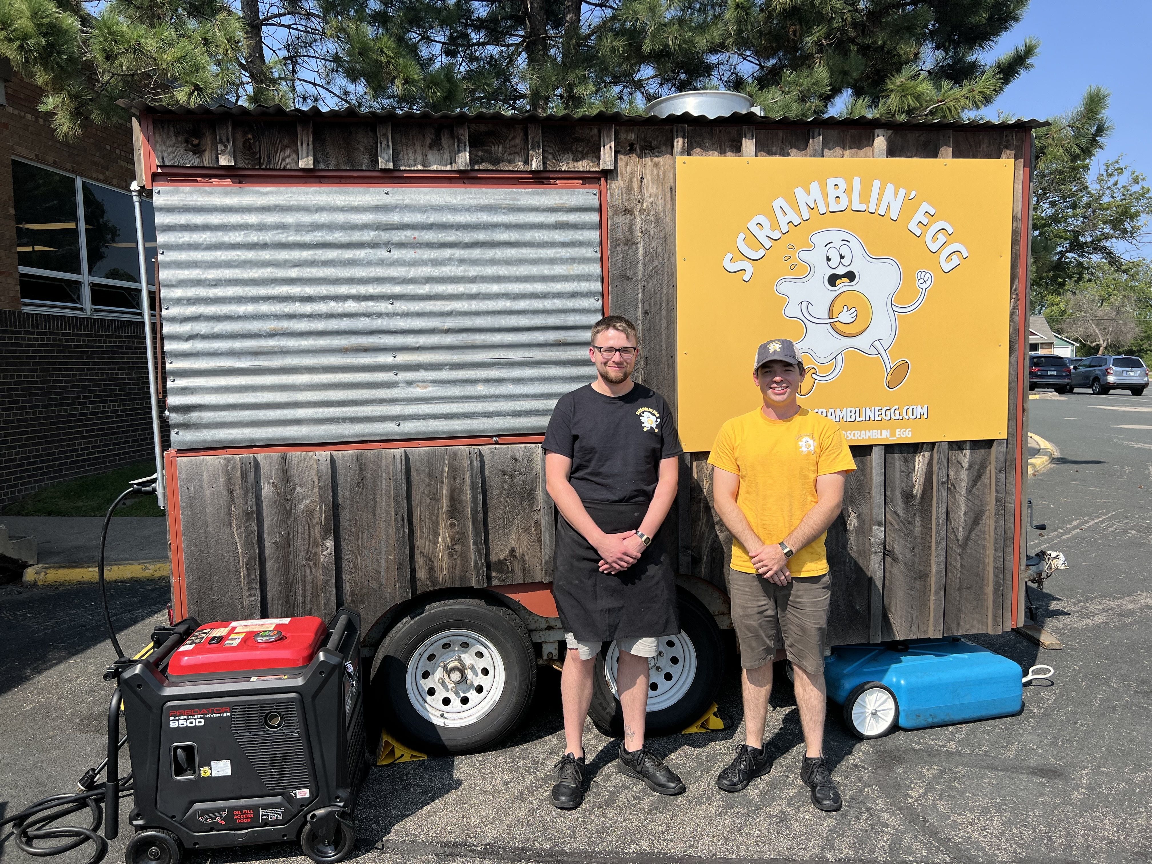 Two men with light skin, one in a black t-shirt and another in a yellow t-shirt, pose in front of a trailer-mounted mobile kitchen, the exterior of which is clad in old weathered barn wood, with an animated egg logo on the side.