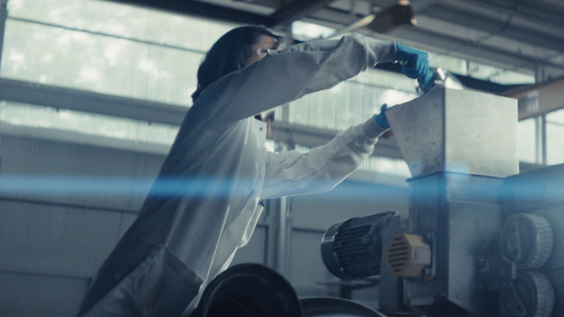 A person in a white lab coat and blue gloves leans over a metal cabinet, reaching toward it in an industrial setting; blurred background shows machinery and a motor.