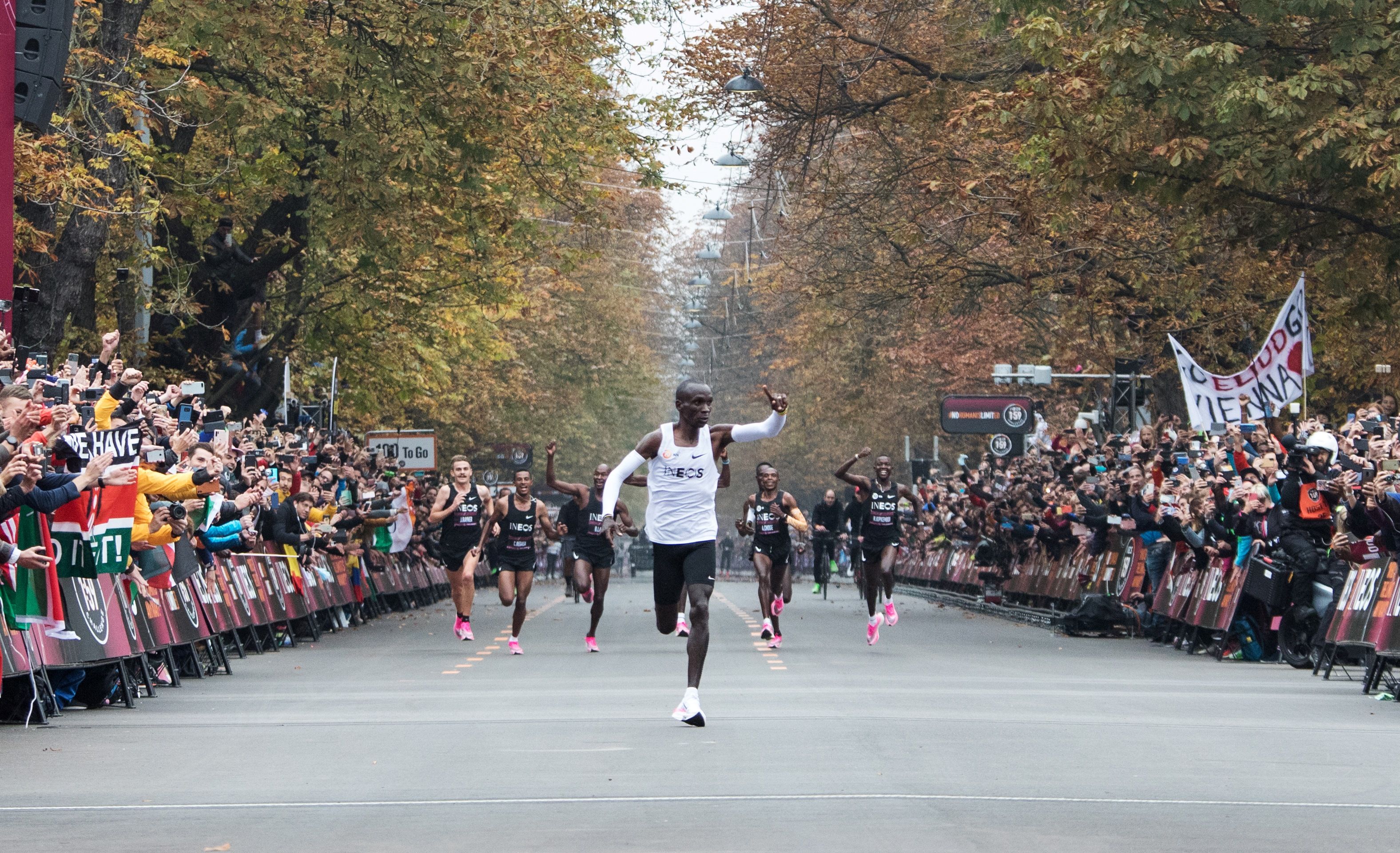 This image is a wideshot of Kipchoge running towards the finish line. Trees and spectators line either side of the road 