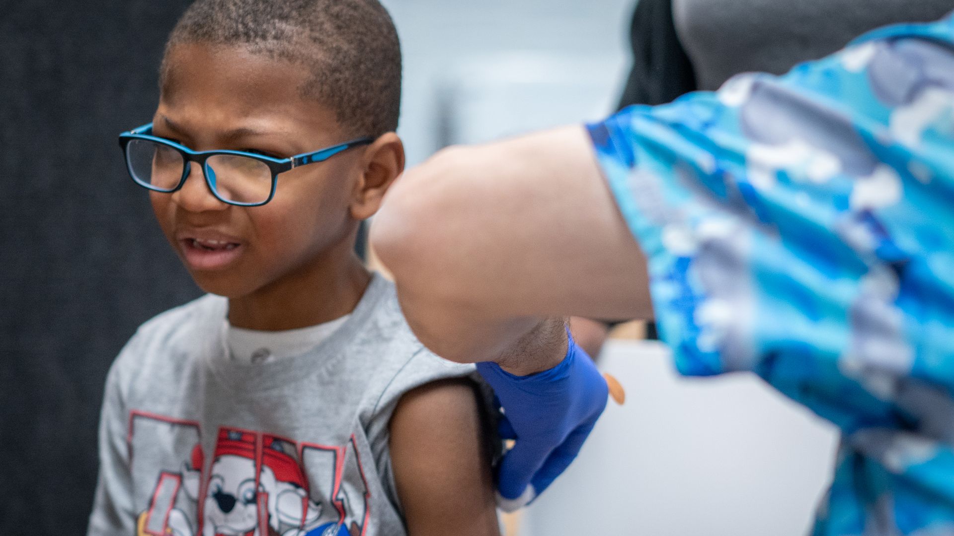 A little boy with glasses gets a measles shot in his left arm.