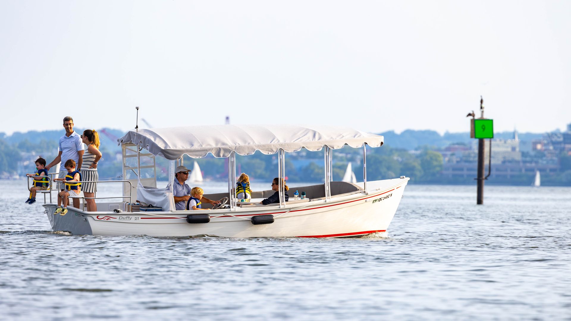 A white boat with a beige canopy carries kids in life jackets and adults on calm water; distant shoreline and a green marker with the number 9 appear in the background.
