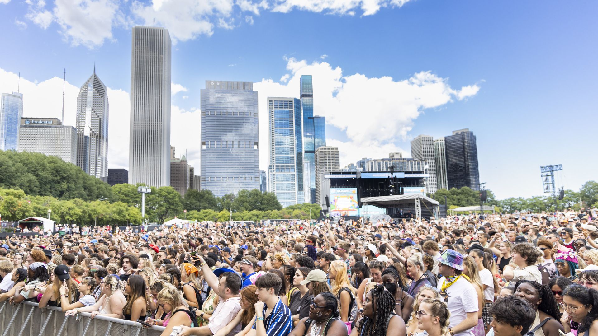 Large diverse crowd at Lollapalooza with Chicago skyscrapers in the background under a partly cloudy blue sky.