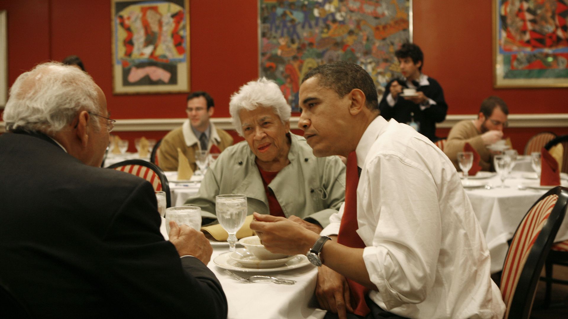 Three people, including chef Leah Chase and President Obama, sit at a table for lunch. Obama is eating something out of a bowl with a spoon.