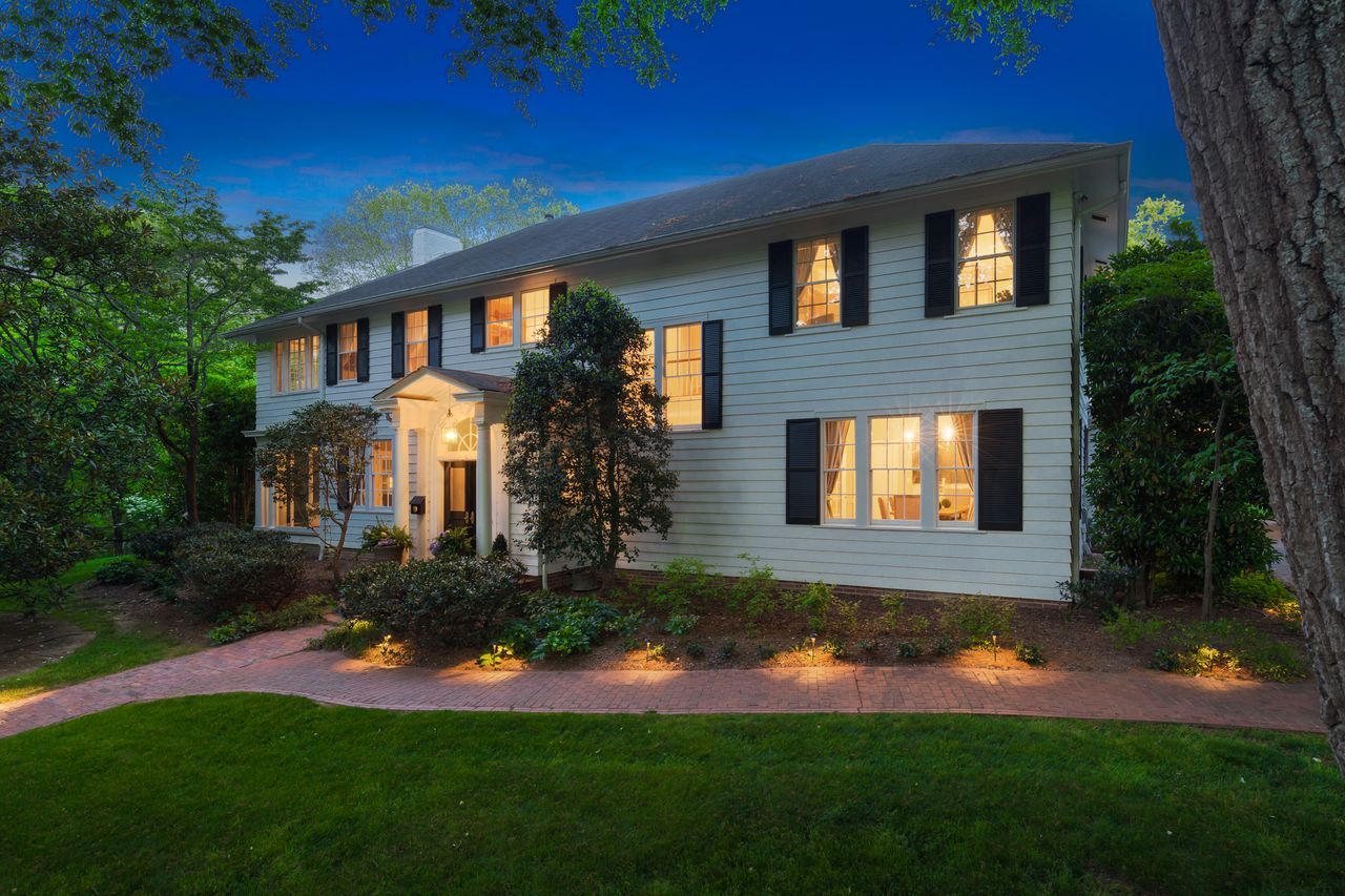 White two-story house with black shutters and warm lights glowing through windows at dusk, surrounded by green trees and a well-lit brick walkway in front.