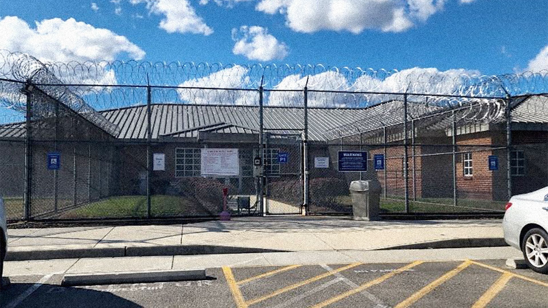 Brick building behind a tall chain-link fence topped with razor wire under a bright blue sky with clouds, with parked cars and sidewalk in the foreground.