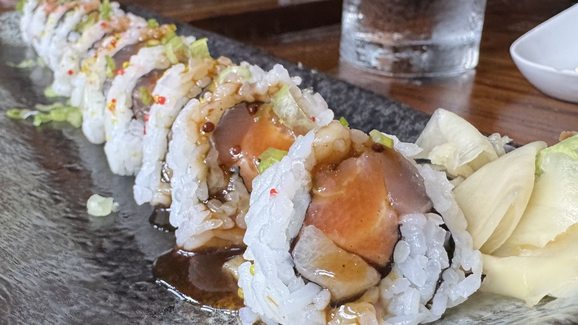 A sushi roll filled tuna and salmon is topped with sauce and green onions, served with wasabi and ginger.