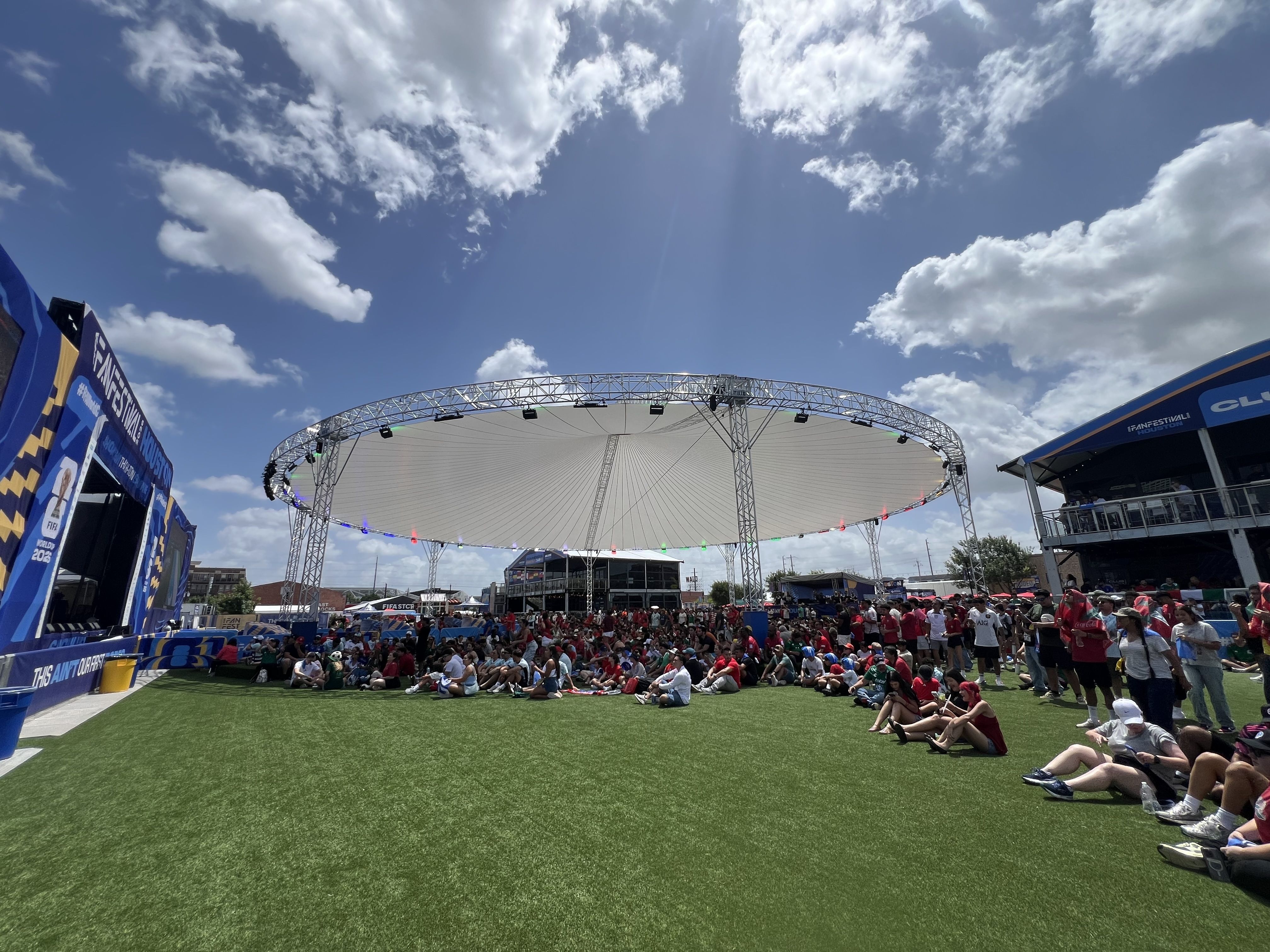 Crowd seated on green artificial turf at a FIFA Fan Festival, facing a large circular stage with white canopy and metal arch; blue and yellow banners frame the area under a bright, partly cloudy sky.