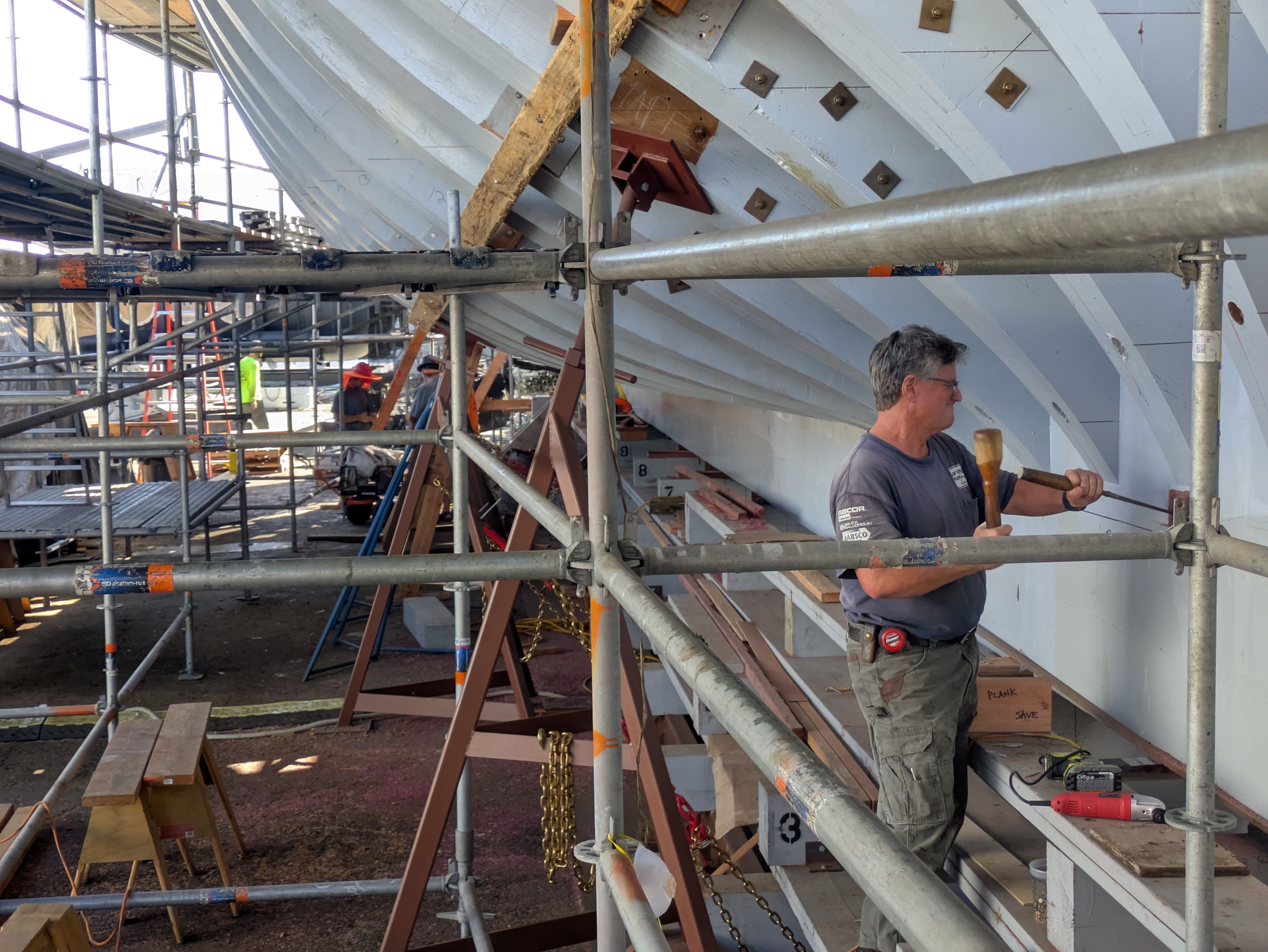 Construction site with metal scaffolding surrounding a large curved light-blue hull under repair. A man in a gray shirt stands on the right holding a tool; workers in hard hats are in the background.