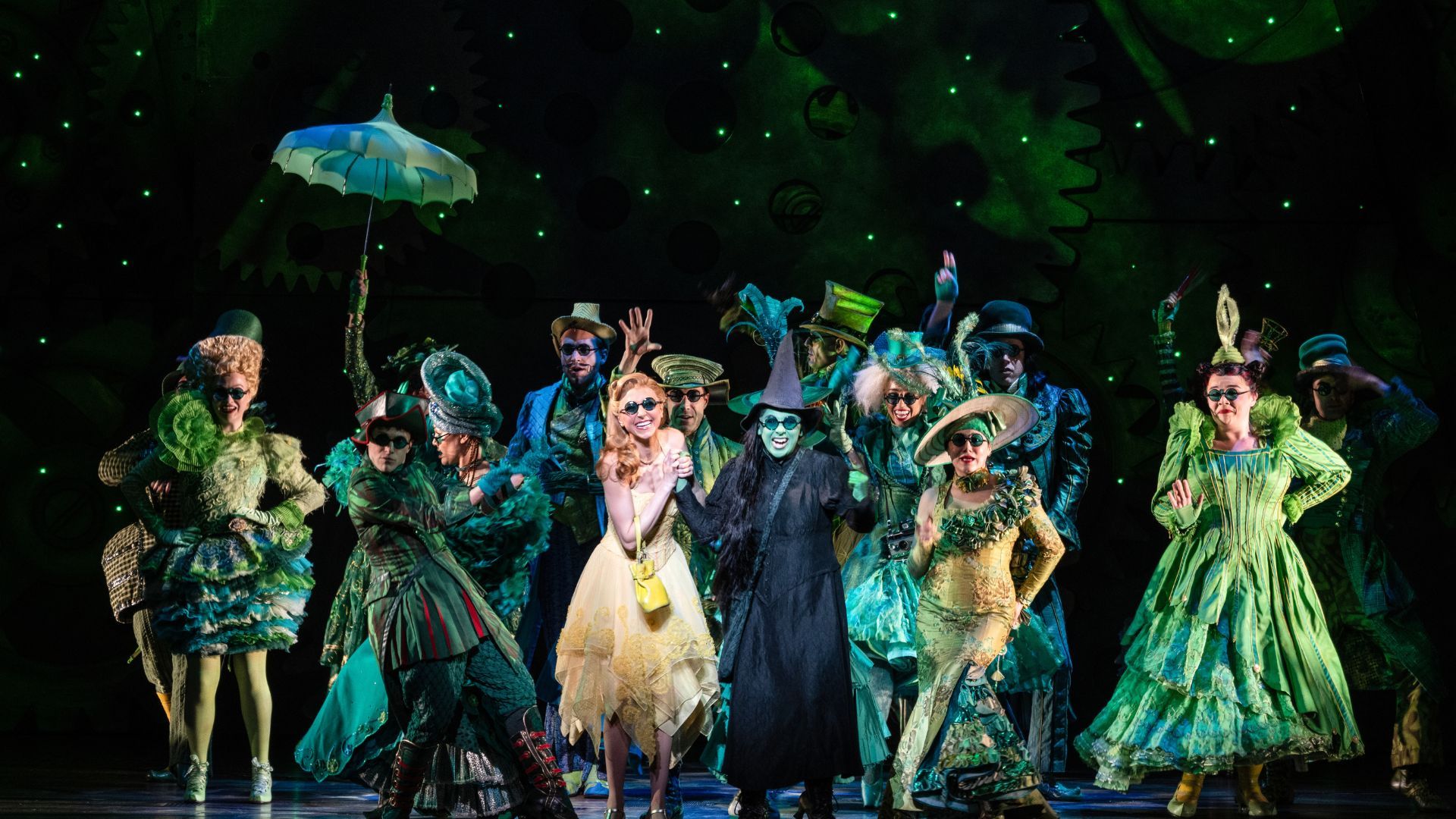 A scene from the musical "Wicked" on a stage. There is a large group of people wearing green clothing.