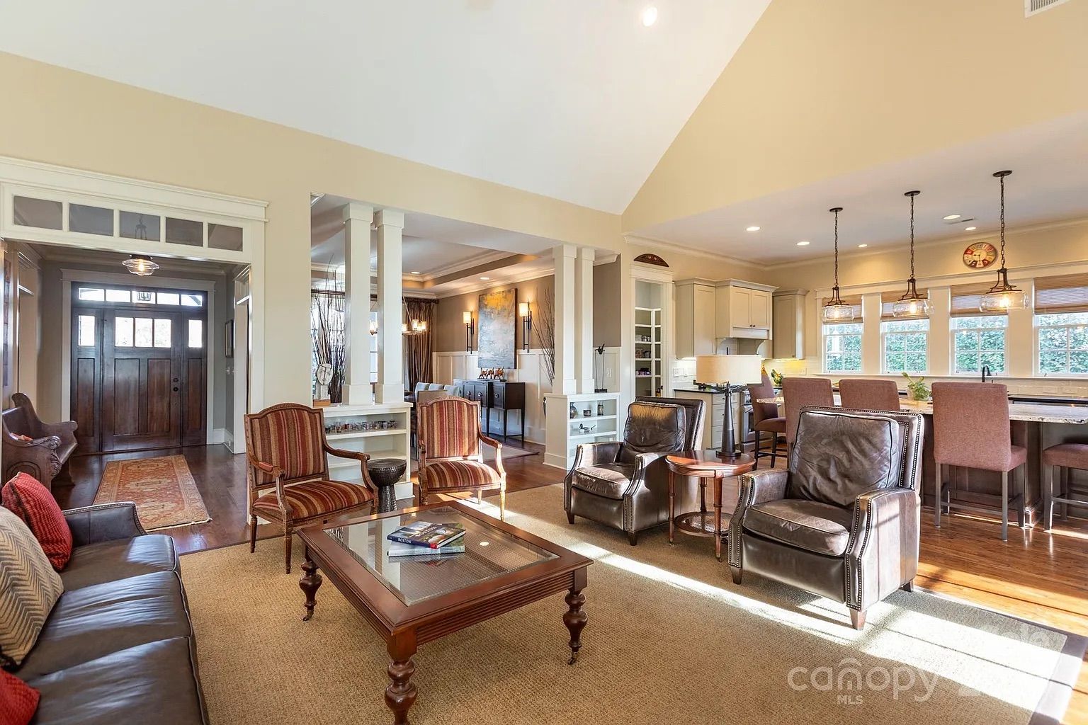 Open living area with beige walls, brown leather sofa, two striped armchairs, two leather armchairs, wooden coffee table, kitchen with beige cabinets, island with bar stools, and pendant lights.