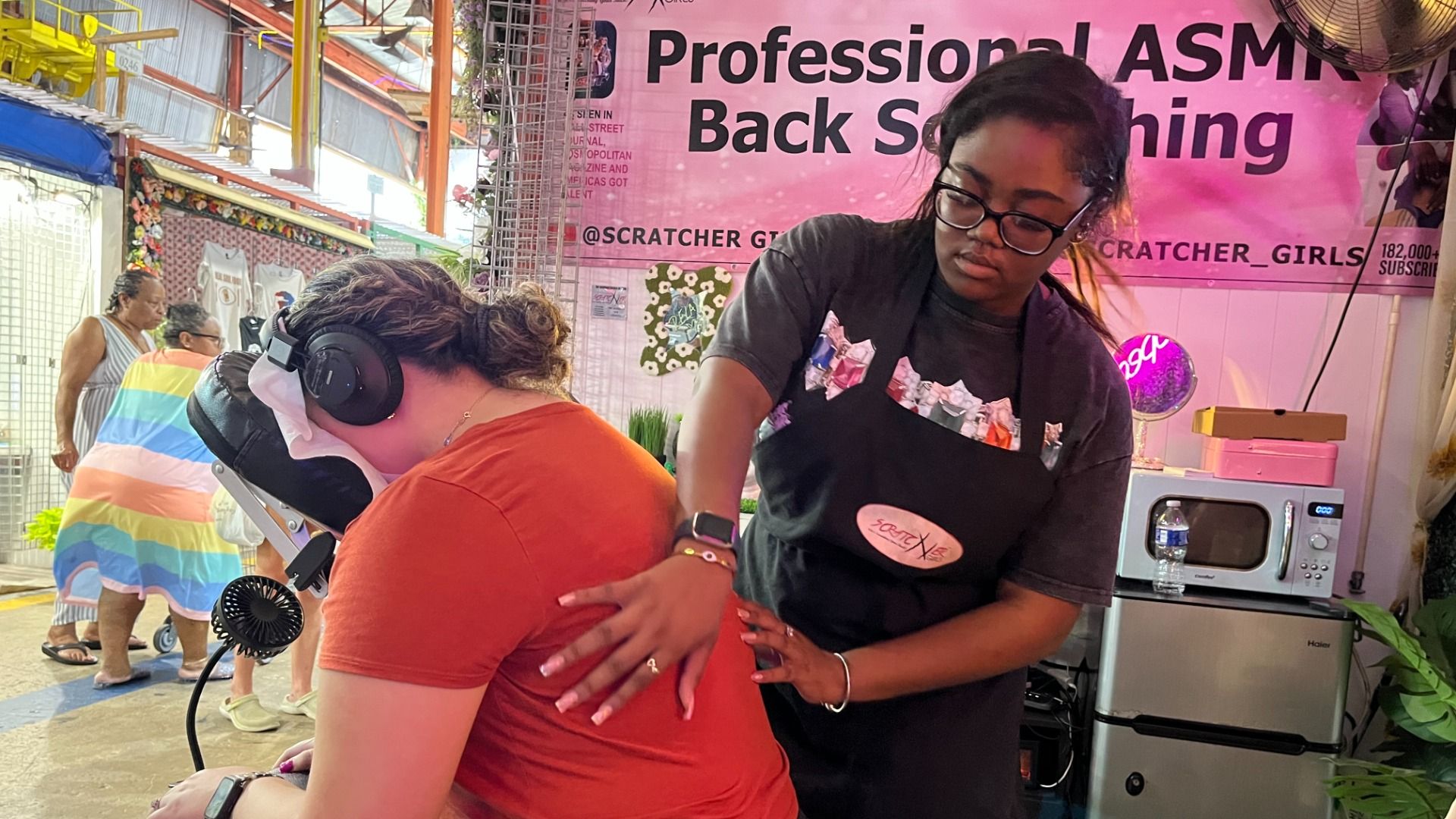 A woman wearing glasses gives a back massage to a seated person in an orange shirt with headphones on, in a market stall with a pink sign reading "Professional ASMR Back Soothing."