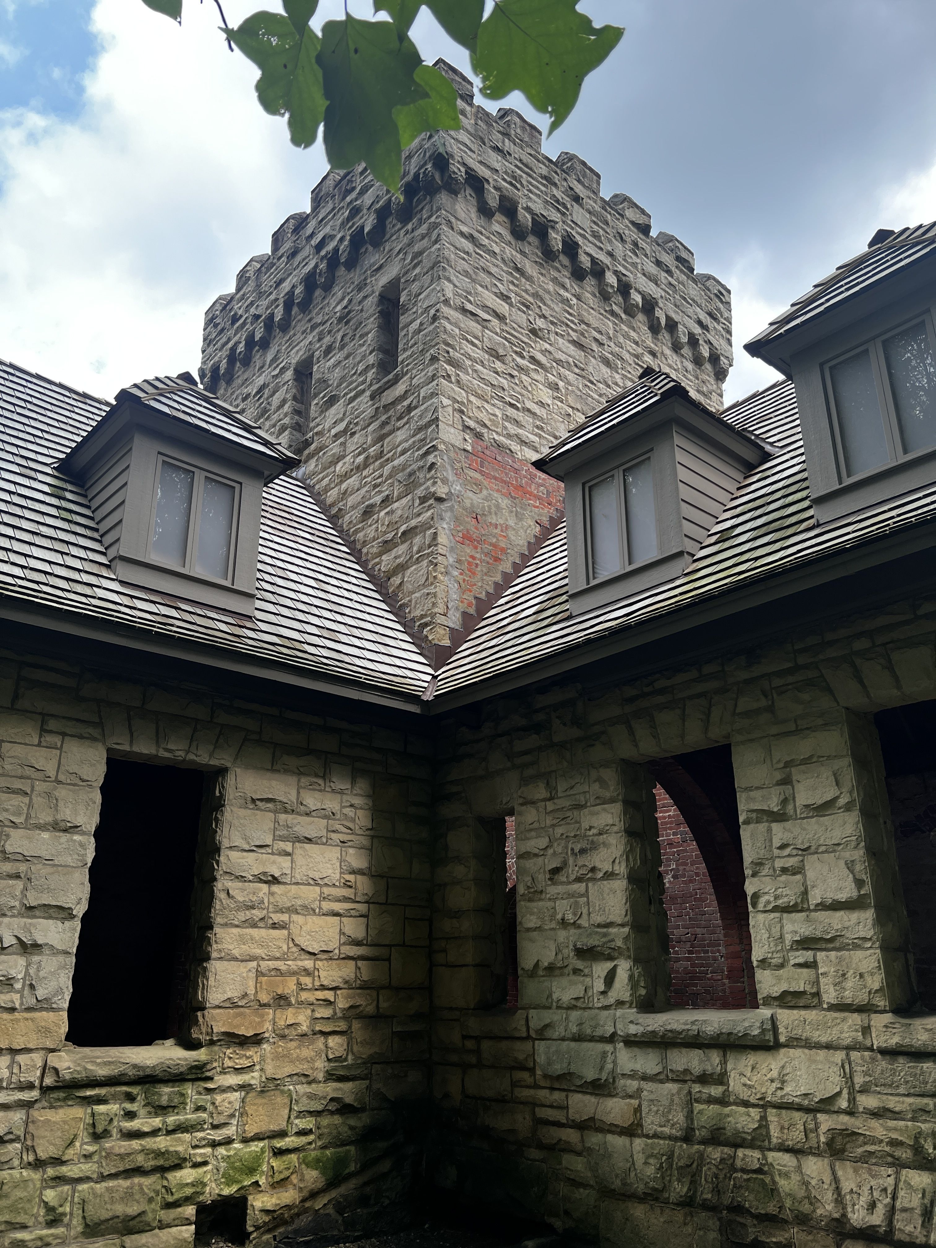 Rugged stone turret with crenellated battlements rises above slate-roofed walls. A red brick section, arched openings, and mossy stonework frame a cloudy blue sky; green leaves hang overhead.