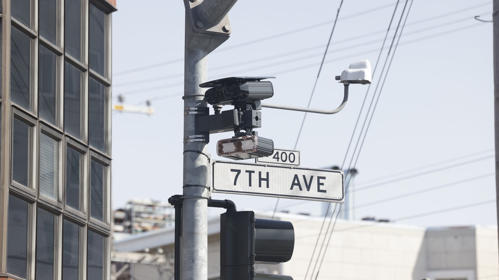 Urban street corner with a metal pole-mounted surveillance camera and a white dome camera on a curved arm, beside a glass-fronted building; street sign reads 7th Ave under a clear blue sky.