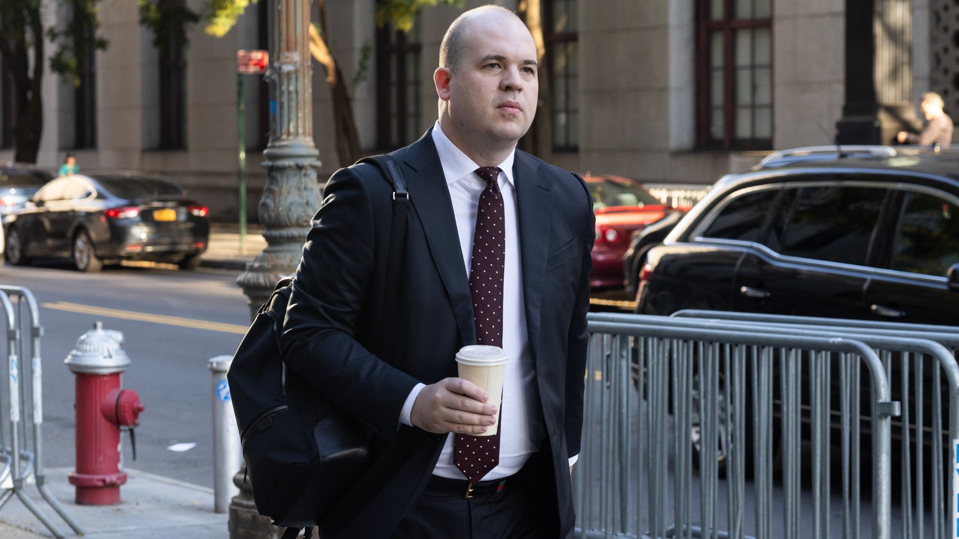 A man in a suit with no hair, walks holding a coffee. 