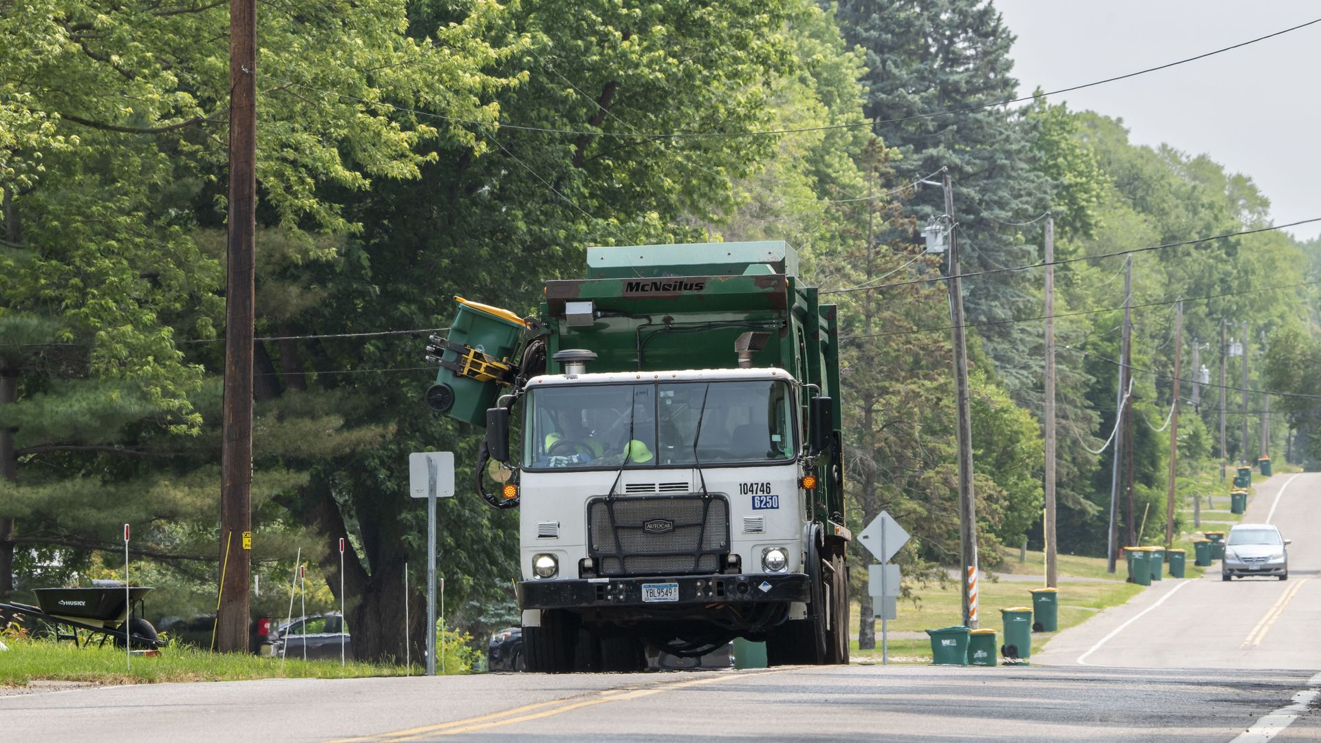 A garbage truck with a white cab and a green bin extends a robotic arm to pick up a green garbage cart along a road with trees in the background.