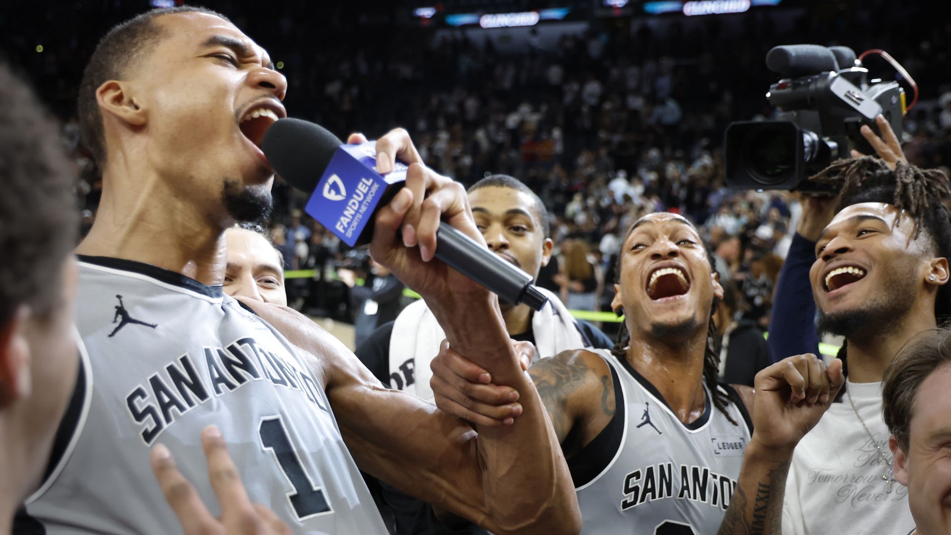 Spurs star Victor Wembanyama yells into a microphone while surrounded by his smiling teammates.