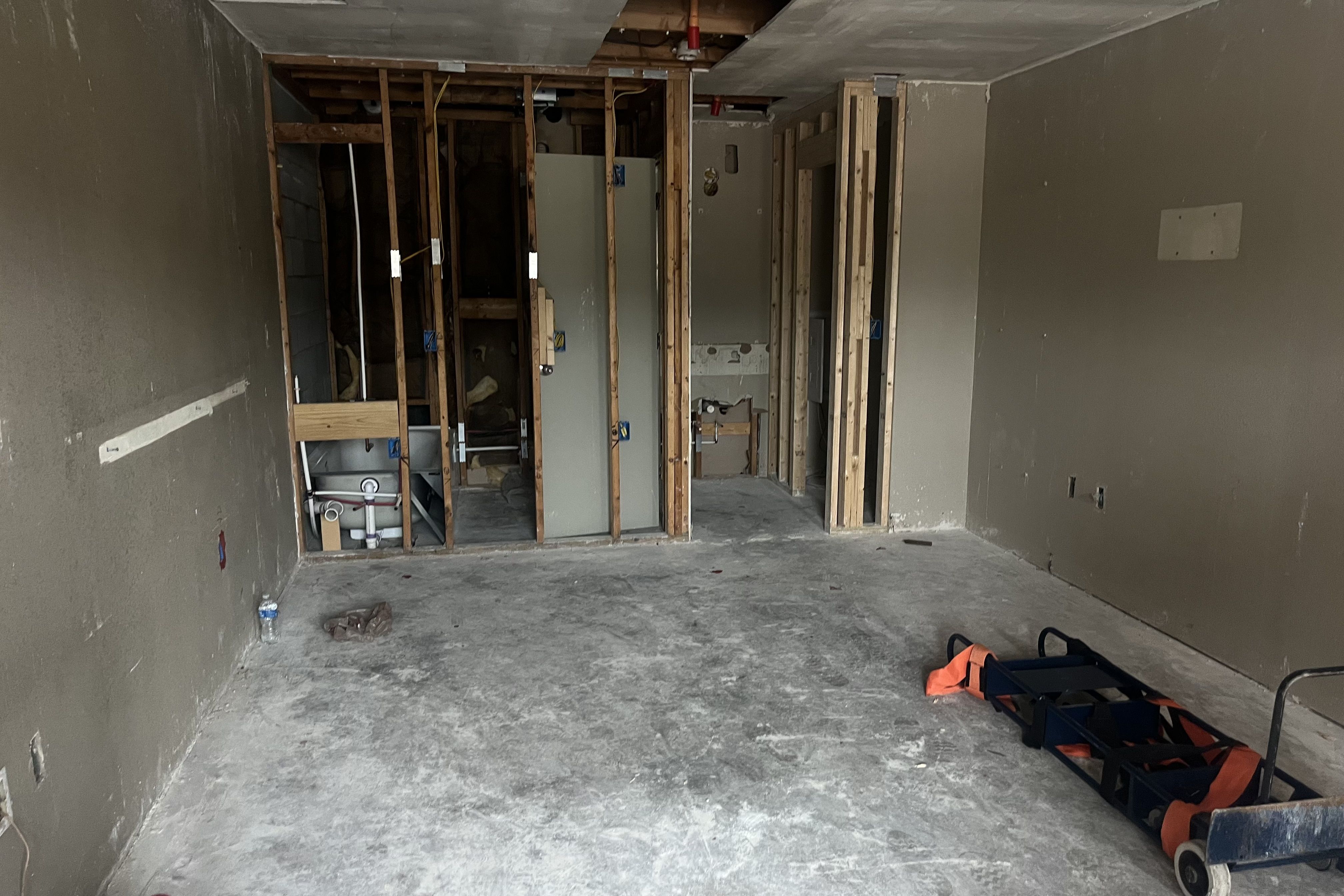 Empty room under renovation with exposed wooden studs, gray concrete floor, gray walls, exposed pipes, and a blue dolly with an orange strap on the right side.