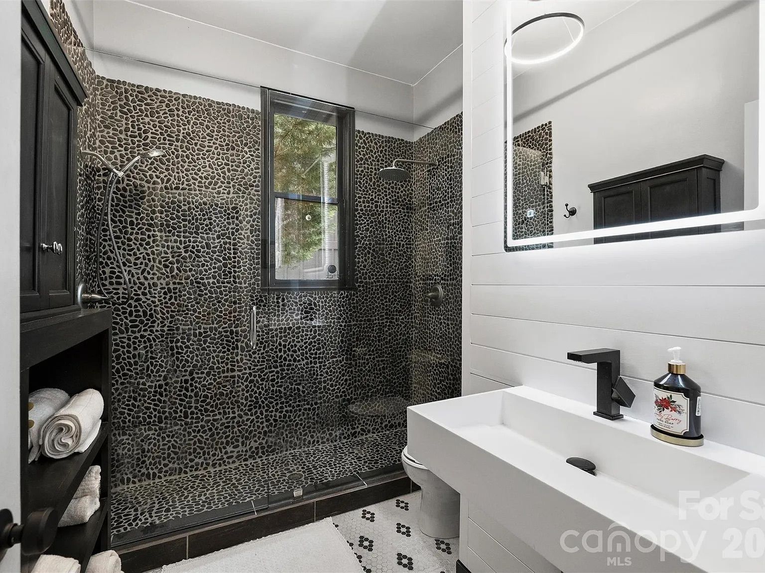 Modern bathroom with a pebble-tiled shower surround in dark tones, a black-framed window, rainfall shower, glass door, white rectangular sink with a black faucet, backlit mirror, black cabinet, and towels.